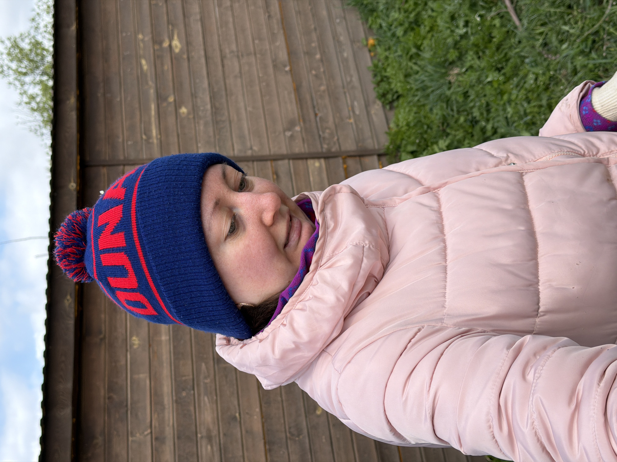 A woman wearing a pink puffer jacket and a blue beanie with a red inscription and pom-pom is pictured outdoors. She is standing near a wooden building or fence, with green grass visible in the background. The photo appears to have been taken in Ol'khovo, Russia.