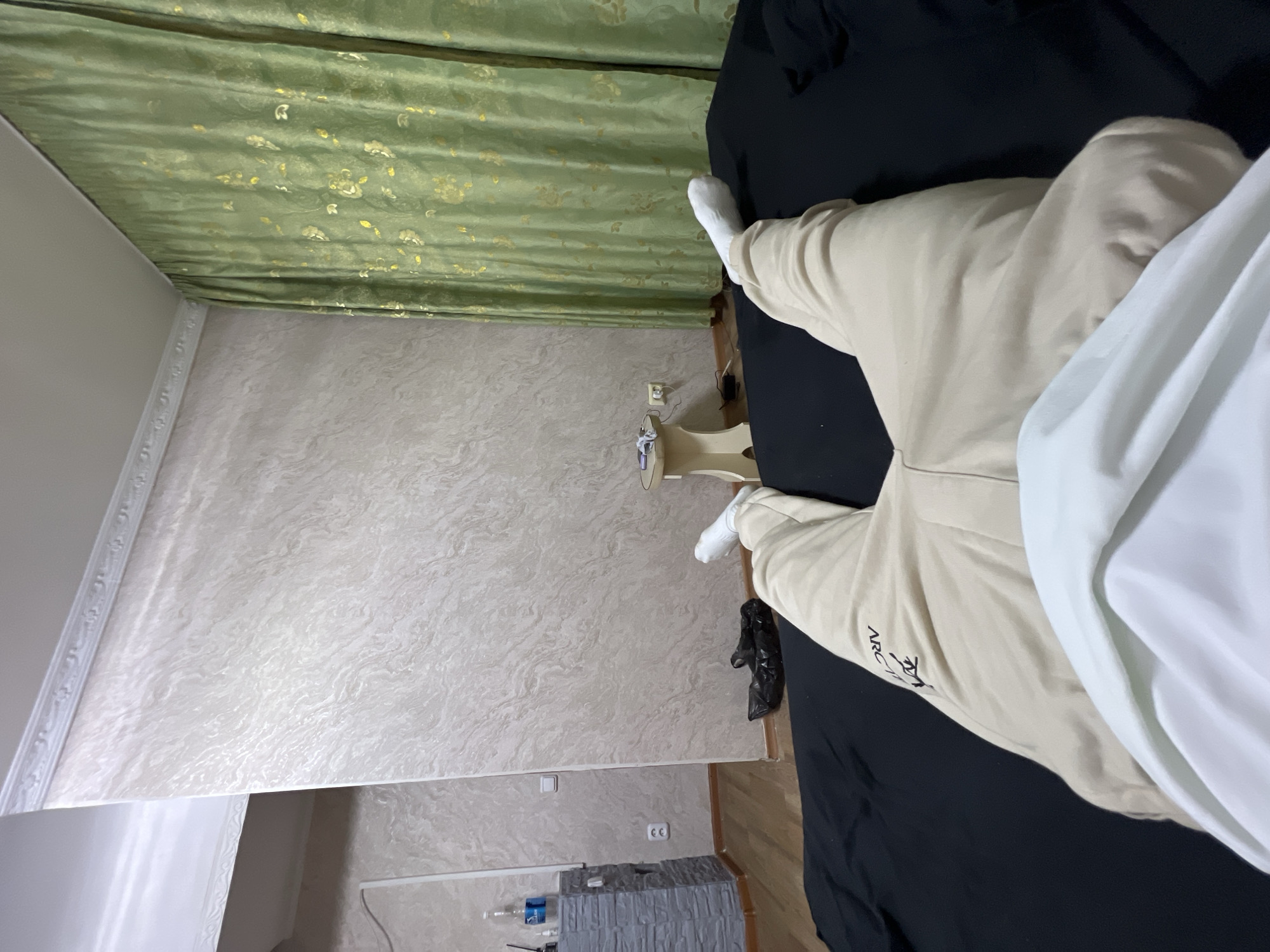 The image shows a person lying on a black surface, presumably a bed, with their legs extended. The person is wearing light-colored sweatpants and white socks. The background features a wall with textured wallpaper in shades of beige and white, and a green curtain with a floral pattern. To the left, a grey dresser-like object with bottles and a black trash bag on the floor are visible. A small, light-colored stool with some items on it is also present. The scene appears to be indoors, possibly a bedroom. The location within the city of Qarağandı city administration, Kazakhstan, is not visually determinable from the image itself.