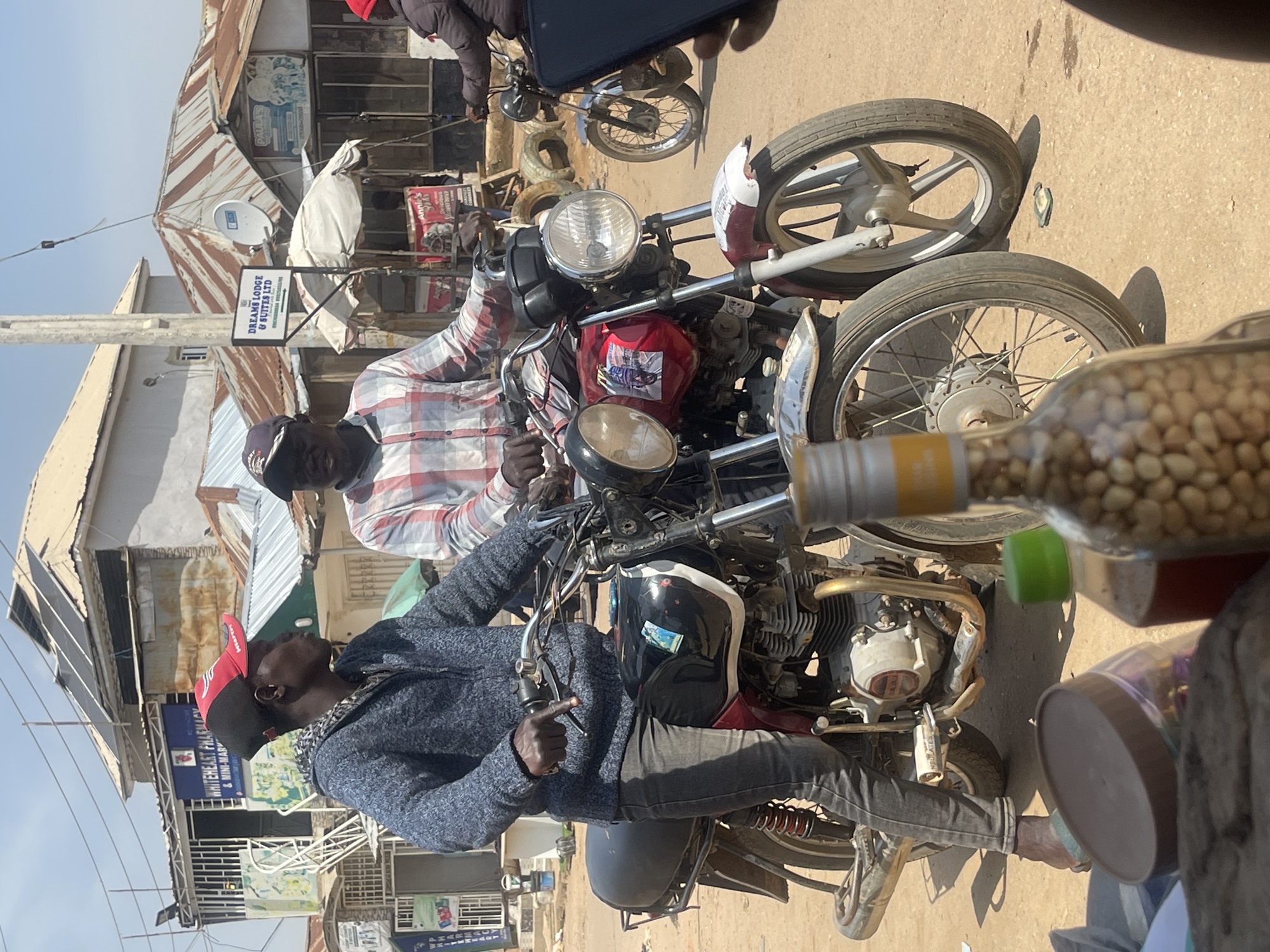 In an outdoor scene in Abuja, Nigeria, two men are positioned on separate motorcycles, likely preparing to depart. The man in the foreground, wearing a grey hooded jacket and a red and black baseball cap, is seated on a black motorcycle and appears to be gesturing with his left hand. To his right, another man in a plaid shirt is on a red motorcycle, his hands on the handlebars. The environment suggests a commercial area with buildings visible in the background, including one with solar panels and another labeled "WHITEHEART PHARMACY & MINI-MART". A sign for "DREAMS LODGE & SUITES LTD" is also present. The ground is dusty and uneven, with some discarded tires visible. In the foreground, partially obscuring the motorcycles, are several bottles and containers, one of which is filled with nuts. The lighting indicates a bright, sunny day.