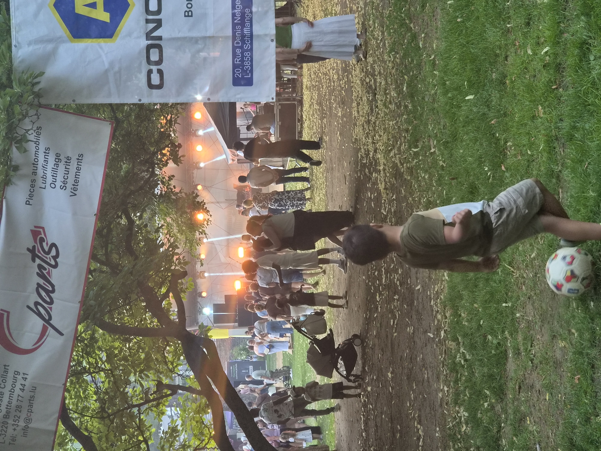 The image depicts an outdoor event with a stage setup in the background, illuminated by stage lights. A crowd of people is gathered, some appearing to be watching a performance or activity. In the foreground, a young boy is playing with a soccer ball. Banners are visible, one displaying the text "parts" and another with an A-shaped logo and the word "CONC". Additional text on the banners indicates sponsorship or informational details, including "L-3220 Bettembourg", "info@cparts.lu", and "20, Rue Denis Netgen L-3858 Schifflange". The setting appears to be a park or public space with trees and grass, with fallen leaves scattered on the ground. The event is likely taking place in or around Differdange, Luxembourg, given the address information present on the banners.