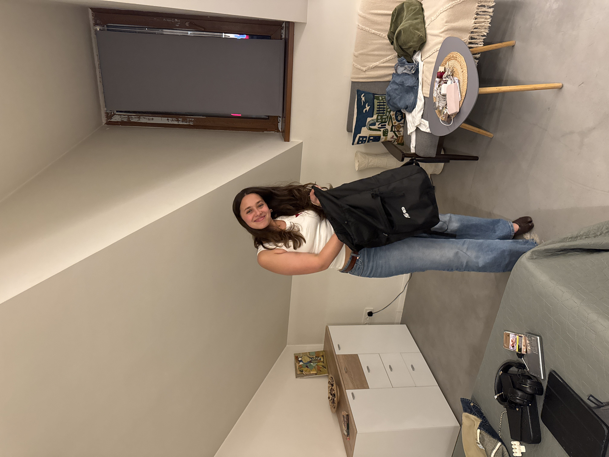 The image displays a young woman standing in an indoor room, likely in Yerevan, Armenia. She has long, dark, wavy hair, and is wearing a white short-sleeved top, blue jeans, and brown shoes. She is smiling cheerfully at the camera, holding a black duffel bag in front of her. The word "Envy" is partially visible on the bag, along with "ED" on another section.
The room features light-colored walls and a sloped ceiling, characteristic of an attic or loft space. A skylight-style window, fitted with a dark roller blind and framed by distressed wood, is prominent at the top. The floor is a smooth, light grey.
To the woman's left, a sofa is adorned with a textured, light-colored throw blanket, a dark green jacket, and some denim clothing. A decorative pillow with a blue and white cityscape pattern rests on the sofa. Adjacent to it is a round grey side table with wooden legs, topped with a woven straw hat containing various small items like jewelry, small bottles, and a pink case.
In the background to the right, a white and light wood dresser or cabinet stands against the wall, with a framed artwork hanging above it. Near the woman's feet, on a grey textured surface (possibly a bedspread or rug), are several electronic devices: a propped-up smartphone, black over-ear headphones, a black power bank, and a black tablet case. The text "E-LIGHT" is visible on a silver charging device or power strip.
The scene conveys a casual and relaxed atmosphere, likely in the evening given the artificial indoor lighting. The woman appears happy and is posing for the photograph.