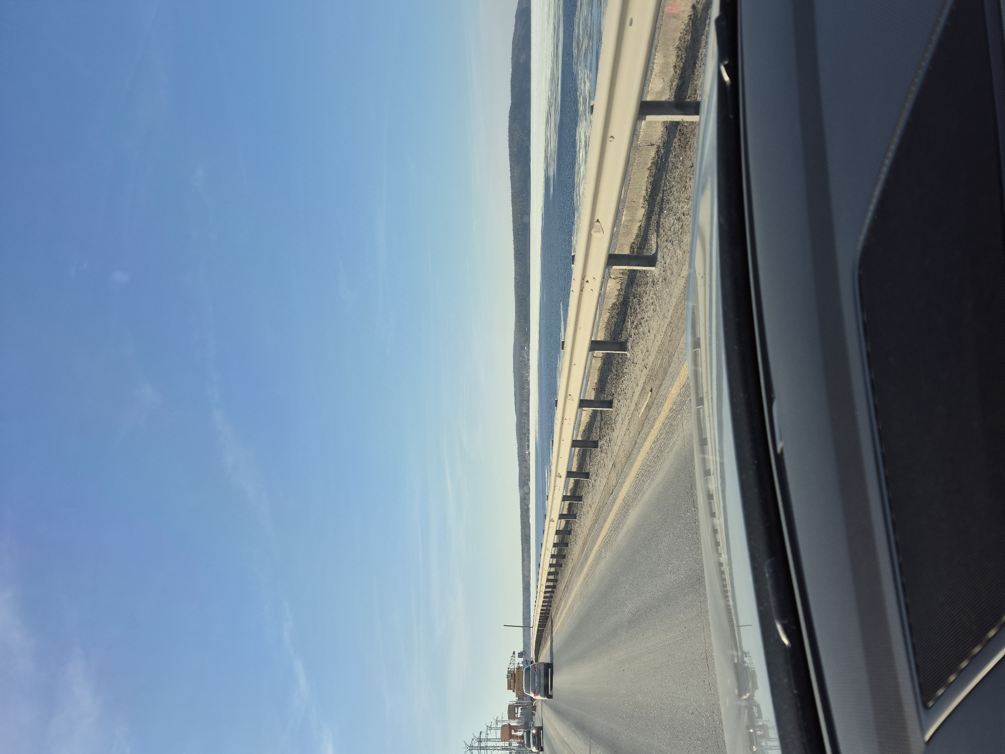 The image depicts a view from inside a vehicle traveling on a highway. A guardrail separates the highway from a body of water, potentially a river or lake, with a strip of land or trees visible on the far side. The sky is clear blue. Traffic is present on the highway. The interior of the vehicle, including a portion of the door panel, is visible. The scene suggests a location near a body of water, likely the Kama River near Chaikovsky, Russia. The photo appears to be taken from a moving vehicle along a highway near the river.