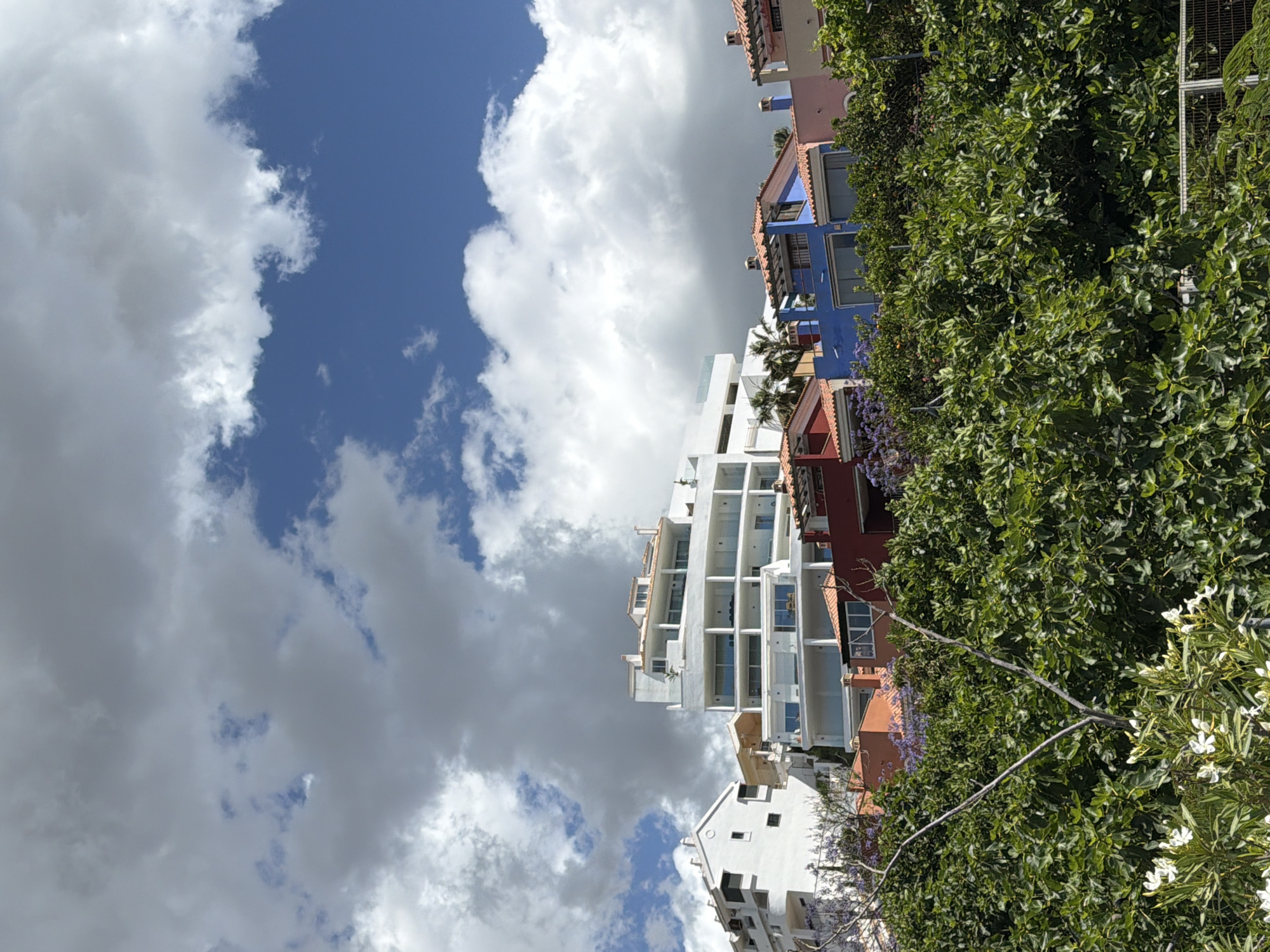 The image depicts a view in Benalmádena, Spain. A series of buildings are visible, including white apartment complexes with balconies and buildings with varied colored facades (blue, pink, red). Lush green trees and foliage are present on the right side of the image. The sky occupies a large portion of the frame, featuring a mix of blue sky and white and gray clouds. The photograph seems to have been taken from a low angle, looking up at the buildings and the sky.