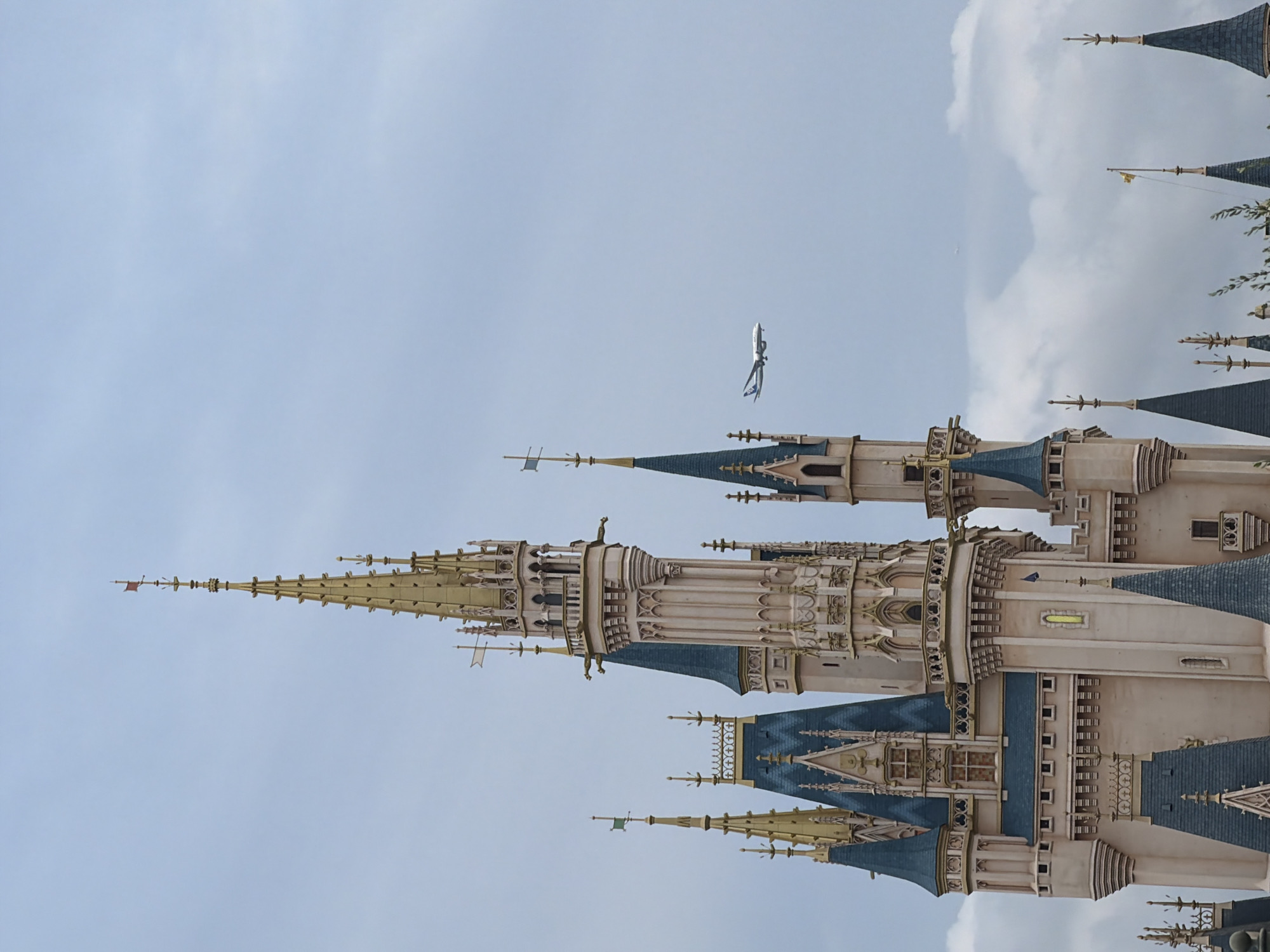 The image shows the Cinderella Castle in Tokyo Disneyland, located in Urayasu, Japan. The castle is a prominent fairytale-inspired structure with multiple turrets and spires, predominantly colored in light beige and accented with gold and dark blue. In the sky to the right of the castle, a commercial airplane is visible flying, its landing gear extended. The sky is overcast with soft grey clouds.