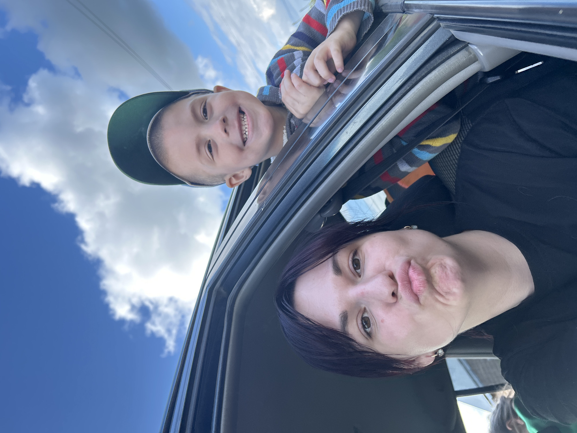 In the city of Fokino, Russia, a selfie is taken from inside a vehicle. Two individuals are visible through the open car window. In the foreground, a woman with dark hair is making a puckering expression. Behind her and to the right, a young boy wearing a green baseball cap and a striped sweater is smiling. The sky, with scattered clouds, serves as the background.
