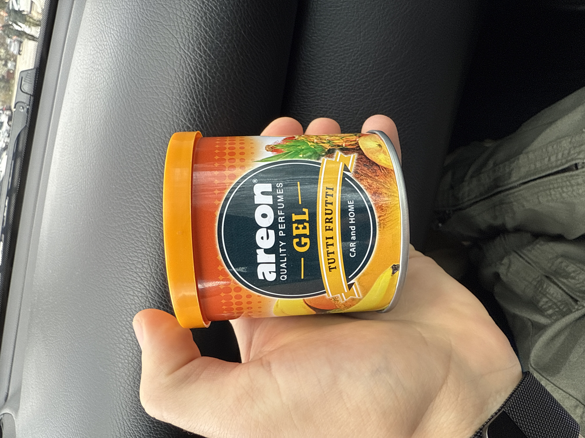 A hand holds an "Areon Tutti Frutti Gel Car and Home" air freshener can. The background shows a car dashboard, black leather car seat, and green pants. The location is not discernible from the image.