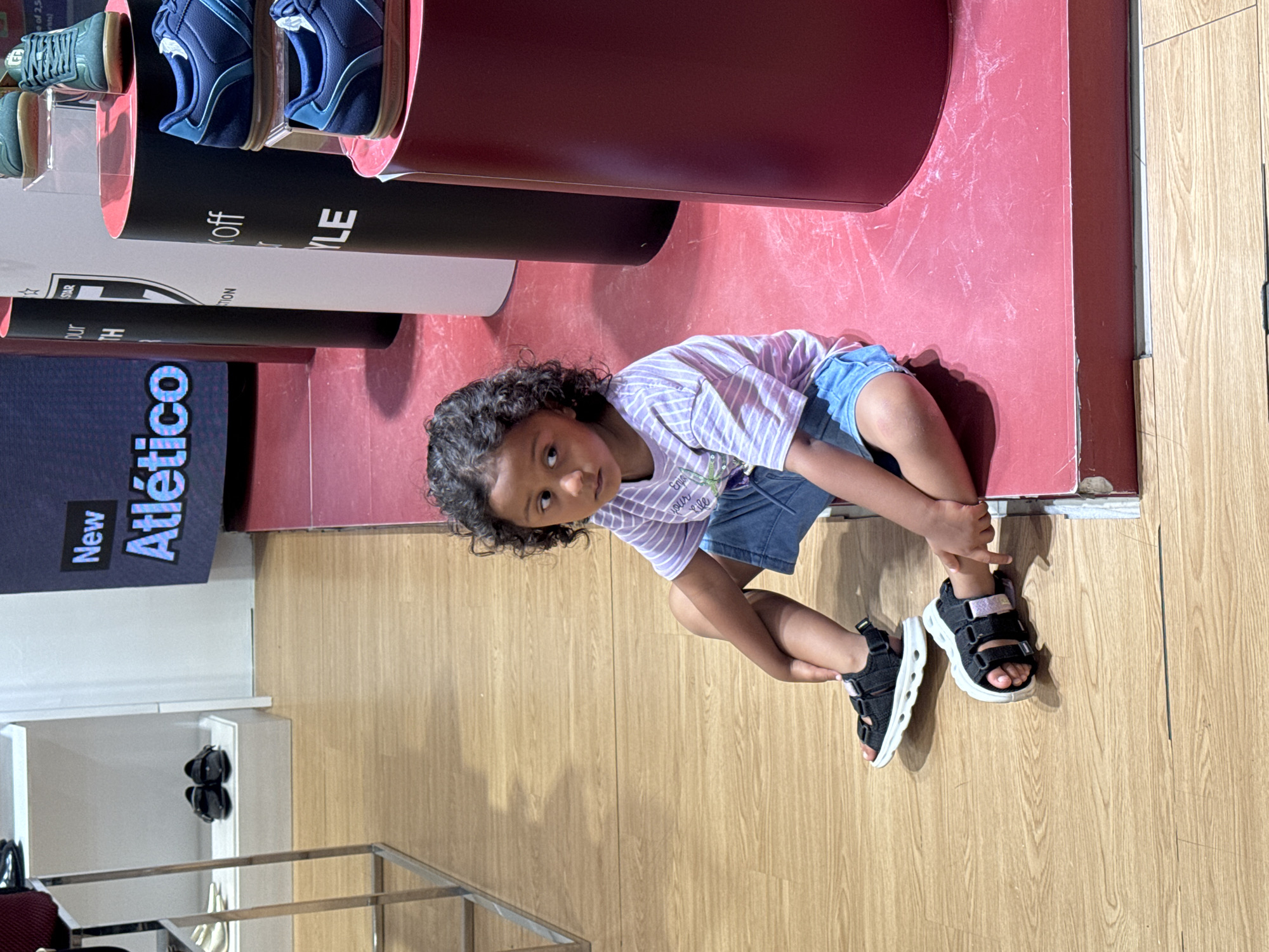A young child with curly dark hair sits on a raised red platform in what appears to be a retail store. The child is wearing a striped purple and white t-shirt and blue denim shorts. They are also wearing black and white sandals with velcro straps. The child is looking to the right of the frame. In the background, there are displays of athletic shoes on red cylindrical pedestals, as well as shelves with more shoes. A large screen displaying text, including the word "New" and "Atlético", is visible in the upper left portion of the image. The floor is made of light-colored wood. The overall setting suggests a shoe store or a retail environment focused on athletic footwear.