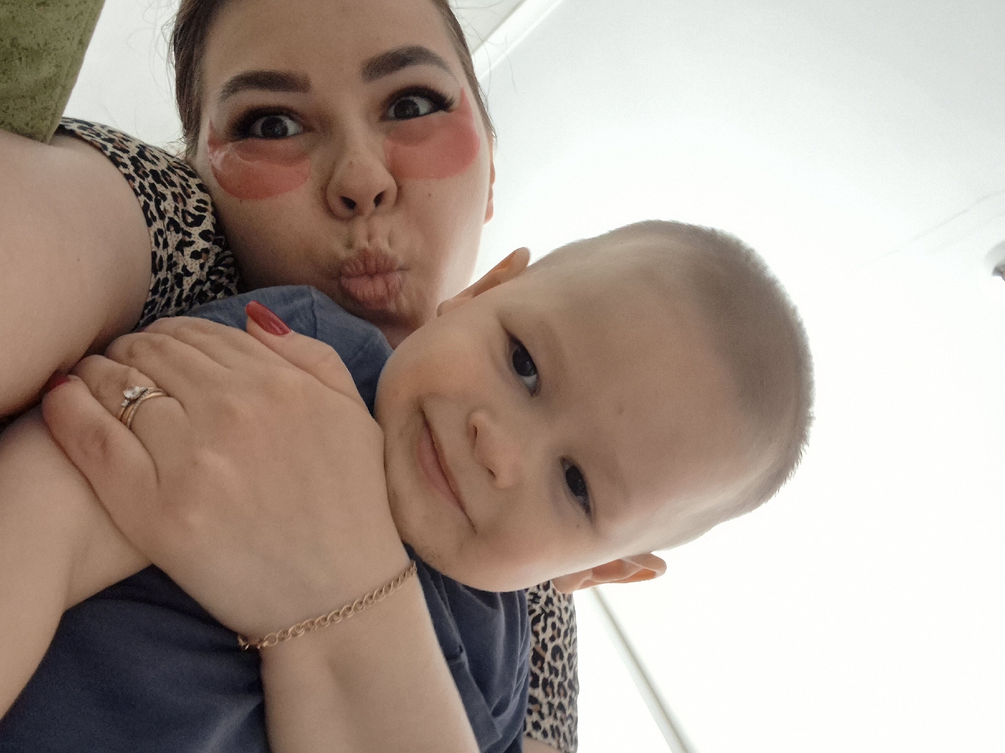 A low-angle, medium shot depicts a woman and a baby. The woman, fair-skinned with dark hair pulled back, wears pink undereye patches. Her mouth is pursed into a kiss. She wears a leopard-print top. Her left hand, with red nail polish, holds the baby. A gold ring is visible on her finger. Her right wrist displays a gold bracelet. The baby, with a bare head, rests in the woman's arms, wearing a blue shirt. The baby looks toward the camera with a slight smile. The background is predominantly white with some green visible near the woman's shoulder. The location is likely indoors, but there are no immediate indications of Tobolsk, Russia.