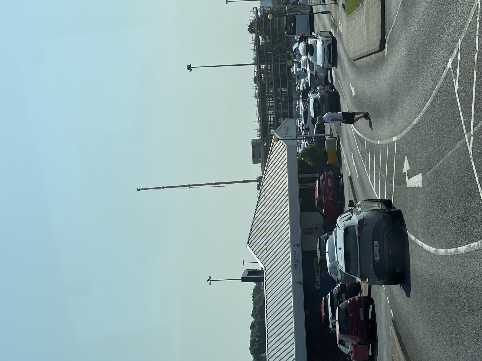 The image shows a sunny day at the Letterkenny Shopping Centre in Letterkenny, Ireland. A white Kia Sportage, license plate 231-DL-1151, is in the foreground, partially obscuring a gray Kia Sportage, license plate 212-LH-7496. To the left, a red Volkswagen Golf, license plate 172-NH-36, is visible. A man in a white t-shirt and black shorts is walking across the road. In the background, there are several cars, a bus, and buildings under construction. The Letterkenny Shopping Centre sign is visible on a white building with a corrugated metal roof.