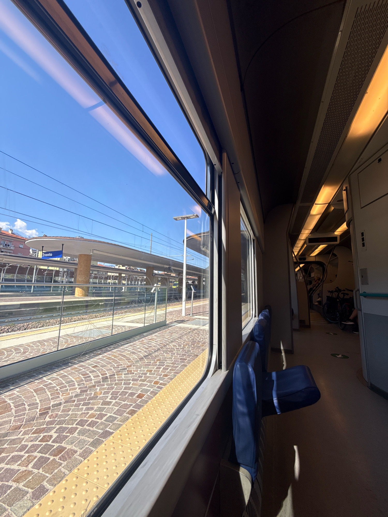 The image shows a view from inside a train carriage looking out of a window. The window is large and reflects the sky and overhead lights. Outside the window, a train station platform is visible, paved with cobblestones and featuring a yellow tactile warning strip. A sign with the word "Trento" in blue lettering hangs above the platform. The station appears to be modern, with a sleek, curved roof structure supported by pillars. In the distance, buildings and power lines are visible under a clear blue sky. Inside the train, blue seats are lined up along the wall, and the corridor stretches ahead, illuminated by warm, overhead lighting. A bicycle is parked in the corridor further down, and a person's legs and feet are visible near it. The overall impression is one of travel and transit in an Italian city.