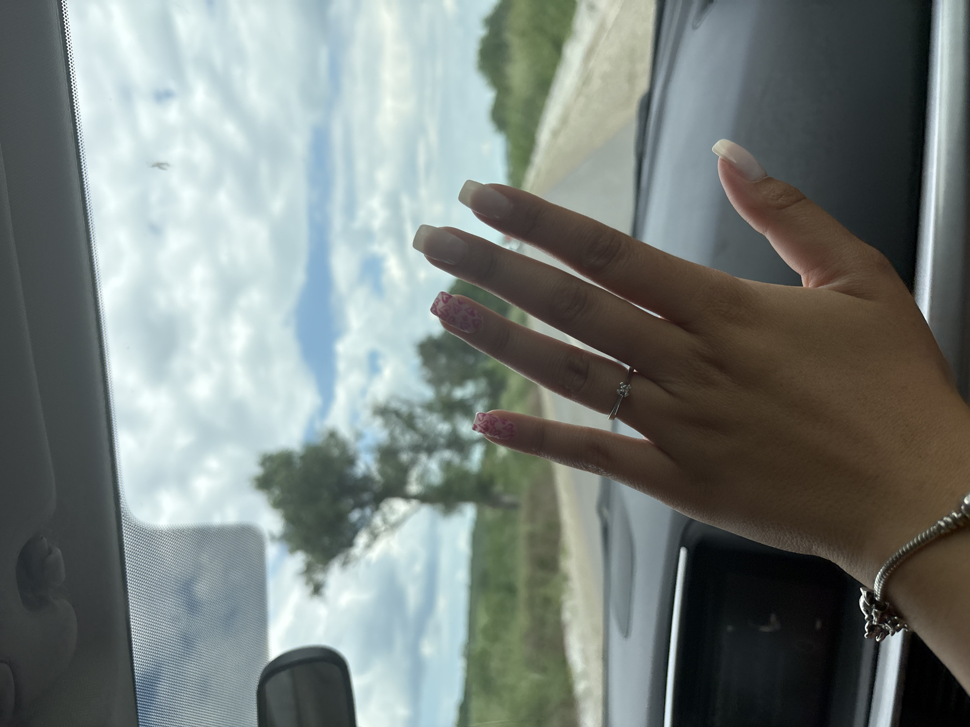A person's hand, adorned with a silver ring on the ring finger and a silver bracelet on the wrist, is positioned against the backdrop of a car windshield. The fingernails are painted in a square-shaped French manicure style, with the ring and pinky fingers displaying a decorative pattern on the tips. The windshield offers a view of a partly cloudy sky and a landscape with trees and a road. The interior of the car is visible, including the dashboard and rear-view mirror. The image appears to be taken from inside the car, facing the windshield, with the person's hand deliberately placed in the foreground. The location of the image is unknown, and there is no information to suggest it is in Dermantsi, Bulgaria.
