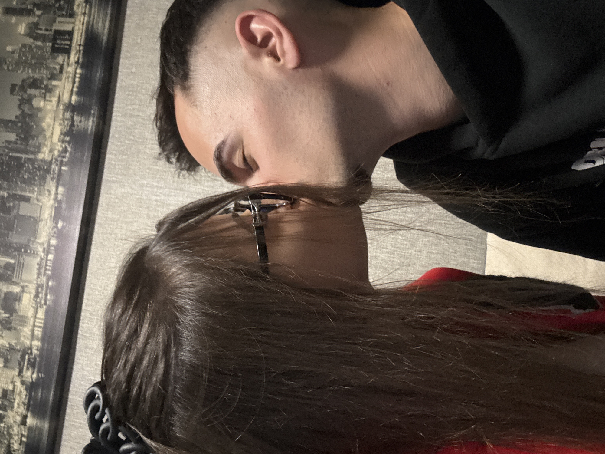 The image depicts two individuals in close proximity. The person on the left has brown hair, is wearing glasses, and appears to be leaning towards the other person. The person on the right has short, dark hair and is wearing a dark-colored hoodie. They appear to be kissing. The background includes a picture frame with a city skyline, and a textured wall. There is no information available in this image to ascertain the location as the city of Sofia, Bulgaria.