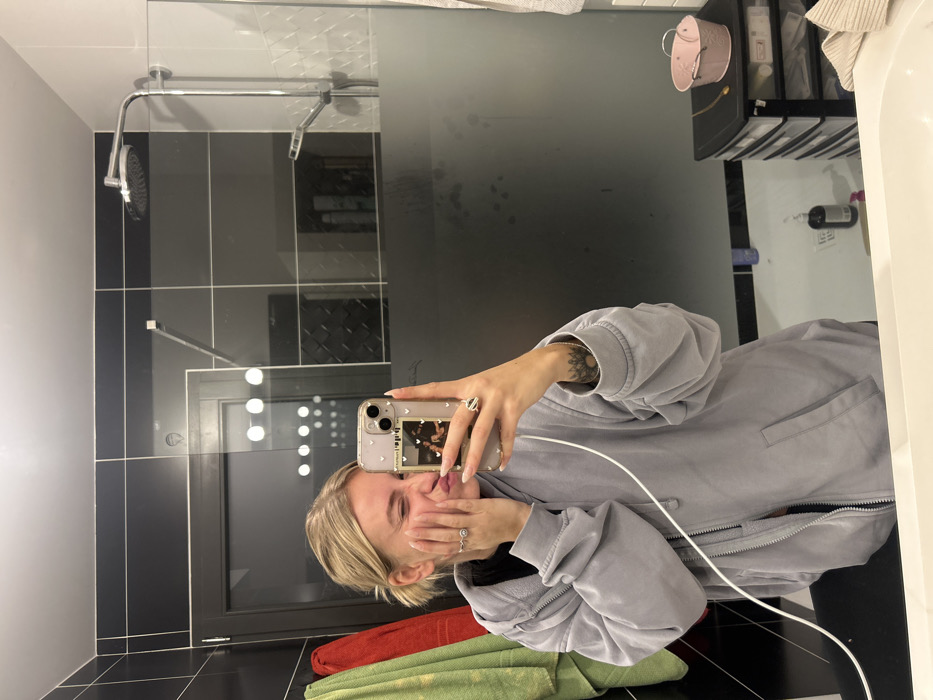 A young woman with blonde hair captures a mirror selfie in a modern bathroom, likely in Chambœuf, France. She is wearing a light grey or lavender-colored hooded jacket with a front zipper. Her left hand, adorned with a ring on the index finger and a visible small tattoo on the forearm, holds a light-colored smartphone with a clear, decorated case that features two small images on the back. A white charging cable is plugged into the phone and drapes across the front of her jacket. Her right hand, with a ring on the ring finger, covers her mouth, suggesting a playful or shy expression with a hint of a smile.
The bathroom features dark grey and black rectangular wall tiles, and darker floor tiles. A clear glass shower screen, partially frosted or steamed in the middle, separates the shower area from the rest of the bathroom. Inside the shower, a metallic shower head and bar are visible. Reflections in the mirror show interior lights and a dark-framed window or door. To the right, a white bathtub or sink edge is visible, along with a small pink bucket hanging on a hook and various personal care bottles on a shelf. Red and green towels are folded on a surface in the lower left foreground. The scene depicts a casual, personal moment.