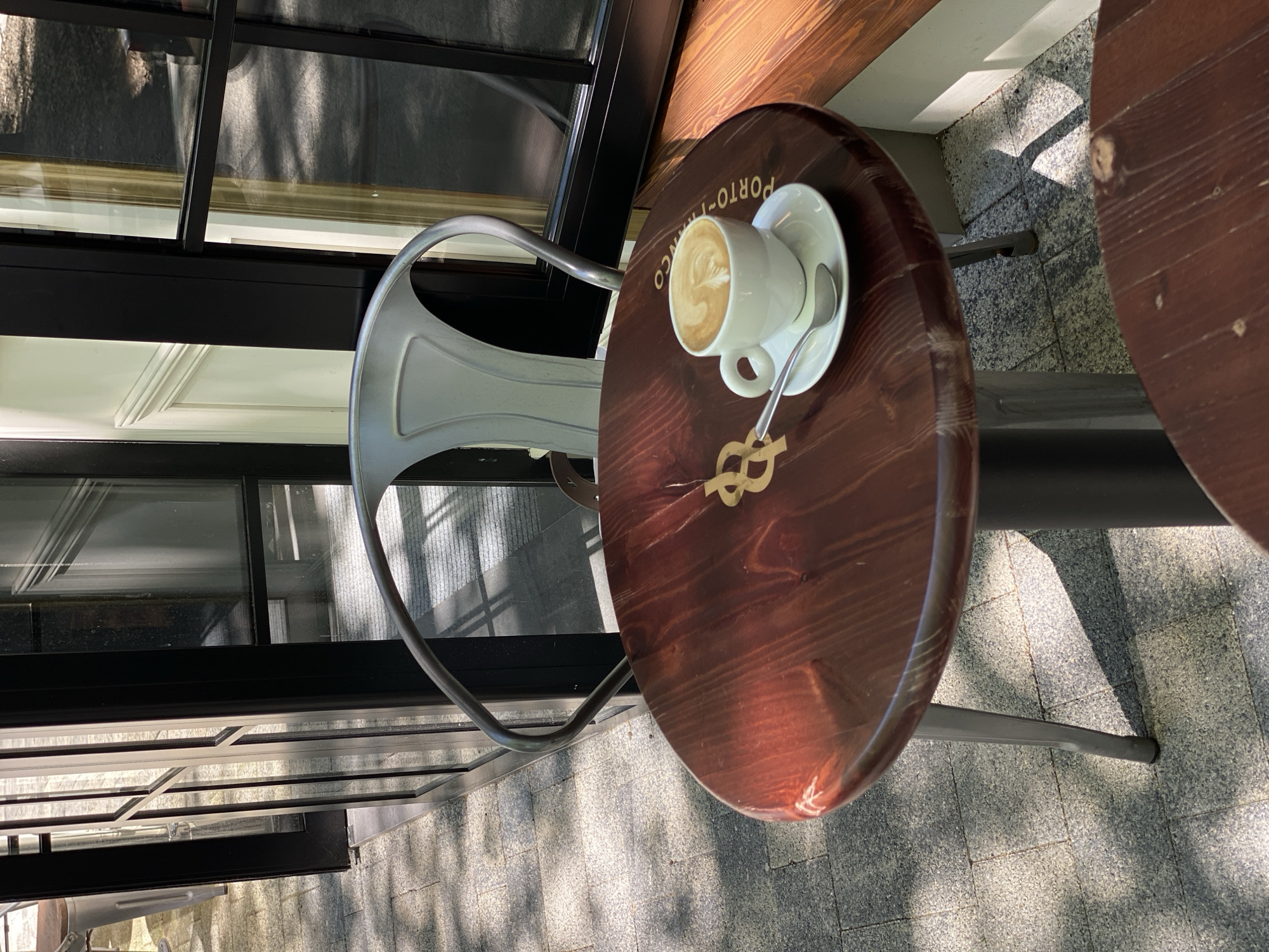 The image displays a serene outdoor café setting in Sochi, Russia, bathed in bright sunlight, indicative of a clear morning or early afternoon. The central focus is a round, dark-stained wooden table. On it rests a white ceramic coffee cup, featuring delicate latte art (likely a heart or rosette design), placed on a matching white saucer with a silver spoon. The table itself is adorned with a golden, stylized knot or figure-eight symbol in its center, and the text "PORTO-FRANCO" is visible along its edge.
Beside the table, a light grey or silver metal chair with a minimalist design awaits a patron. A second, identical wooden table is partially visible to the right, suggesting multiple outdoor seating arrangements. The tables and chair are situated on a paved surface, which appears to be composed of light grey granite cobblestones or square tiles. Strong sunlight casts distinct, elongated shadows across this ground.
In the background, a building features large, dark-framed windows that reflect parts of the sky and perhaps some surrounding greenery or trees. Above these windows, there is warm-toned wooden paneling, possibly forming part of the building's facade or an awning structure. No people are present, lending a quiet and inviting atmosphere to the scene, suggesting a peaceful moment for a coffee break in a pleasant outdoor environment.
