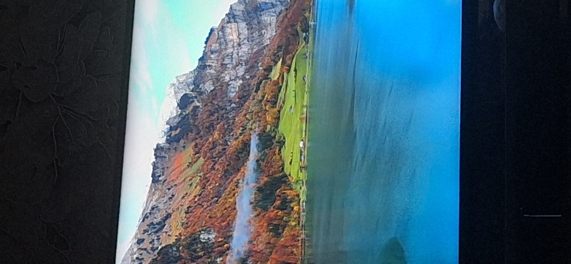 This image depicts a scenic mountain landscape with a body of water in the foreground. The mountains are covered with trees displaying autumn foliage in various shades of orange, red, and yellow, with some patches of green. The rocky peaks of the mountains are partially covered in snow. A light mist or fog is visible near the base of the mountains, partially obscuring the trees. In the middle ground, there are some green fields with a few small buildings that appear to be houses or barns. The foreground is dominated by a large, calm body of water, likely a lake or a wide river, which reflects the colors of the sky and mountains. The water transitions from a deep blue in the lower portion of the image to a lighter turquoise near the shore. The sky is a clear, pale blue. The image appears to be a photograph taken of a screen, indicated by the dark, textured border at the top and bottom of the frame, suggesting it's a display of some sort. The location of Bila Tserkva, Ukraine is not depicted or suggested in this image, as the scene is mountainous and characterized by a large body of water.