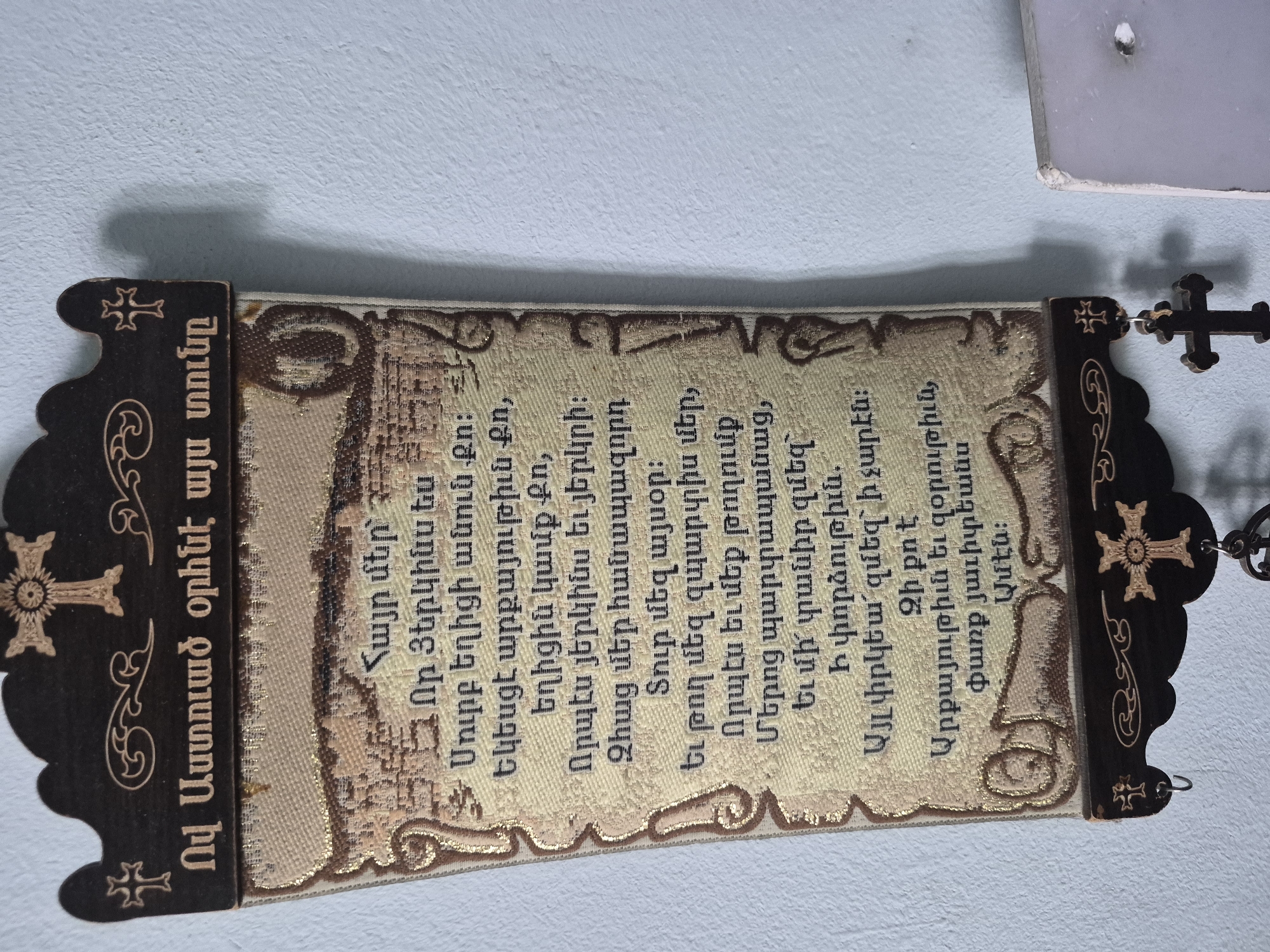 The image shows a close-up of an Armenian religious text, likely a prayer or scripture, displayed on a wall. The text is written in Armenian script on a fabric material with ornate gold trim. The fabric is attached to a dark wooden plaque at the top, which features decorative carvings and crosses. Below the fabric text, the wooden plaque continues downwards and has a cross with a hanging wooden cross suspended from it. The background is a light blue, textured wall, typical of interior settings. The overall impression is of a devotional item displayed within a home or religious space in Yerevan, Armenia.