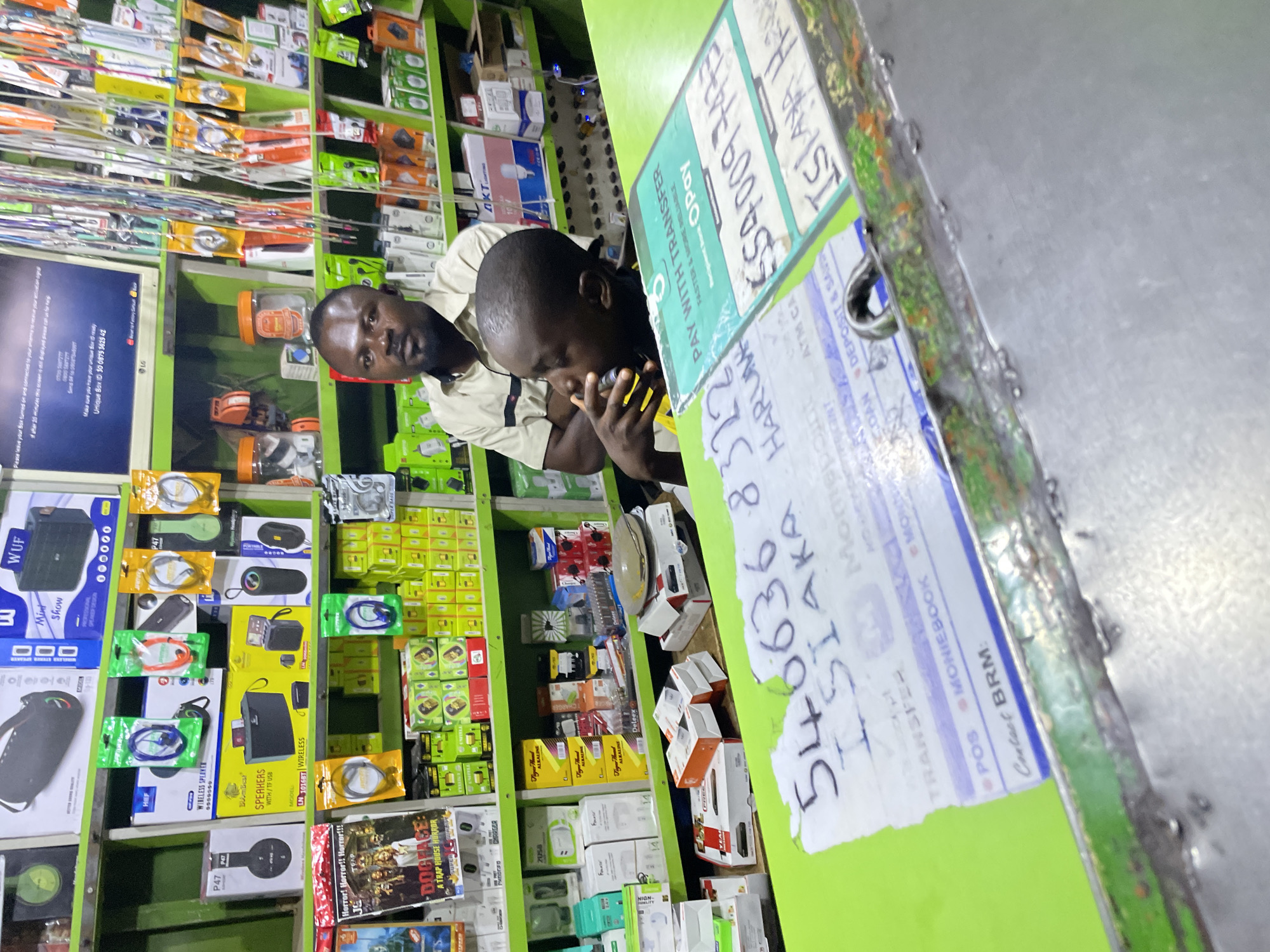 The image depicts the interior of a small retail store, likely in Jos, Nigeria, with shelves packed with electronics accessories, speakers, chargers, and batteries. Two dark-skinned men are present; one is closer to the camera, holding a cell phone to his ear. The other man stands behind him, partially obscured. A green counter is visible in the foreground, covered in printed receipts or labels with handwritten numbers on them. A computer monitor and a variety of electronic products line the shelves.