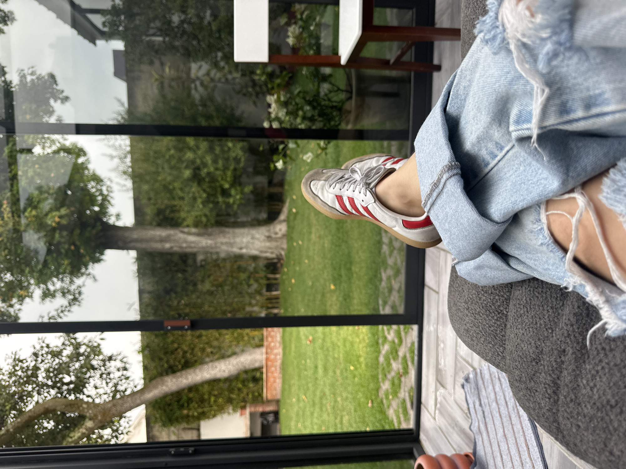 A person sits indoors, one leg crossed over the other, wearing distressed denim jeans and white sneakers with red stripes. Through the window behind them, a green lawn and trees are visible. The location is likely Naucalpan de Juárez, Mexico. A portion of a white chair and wooden stool are also visible indoors.