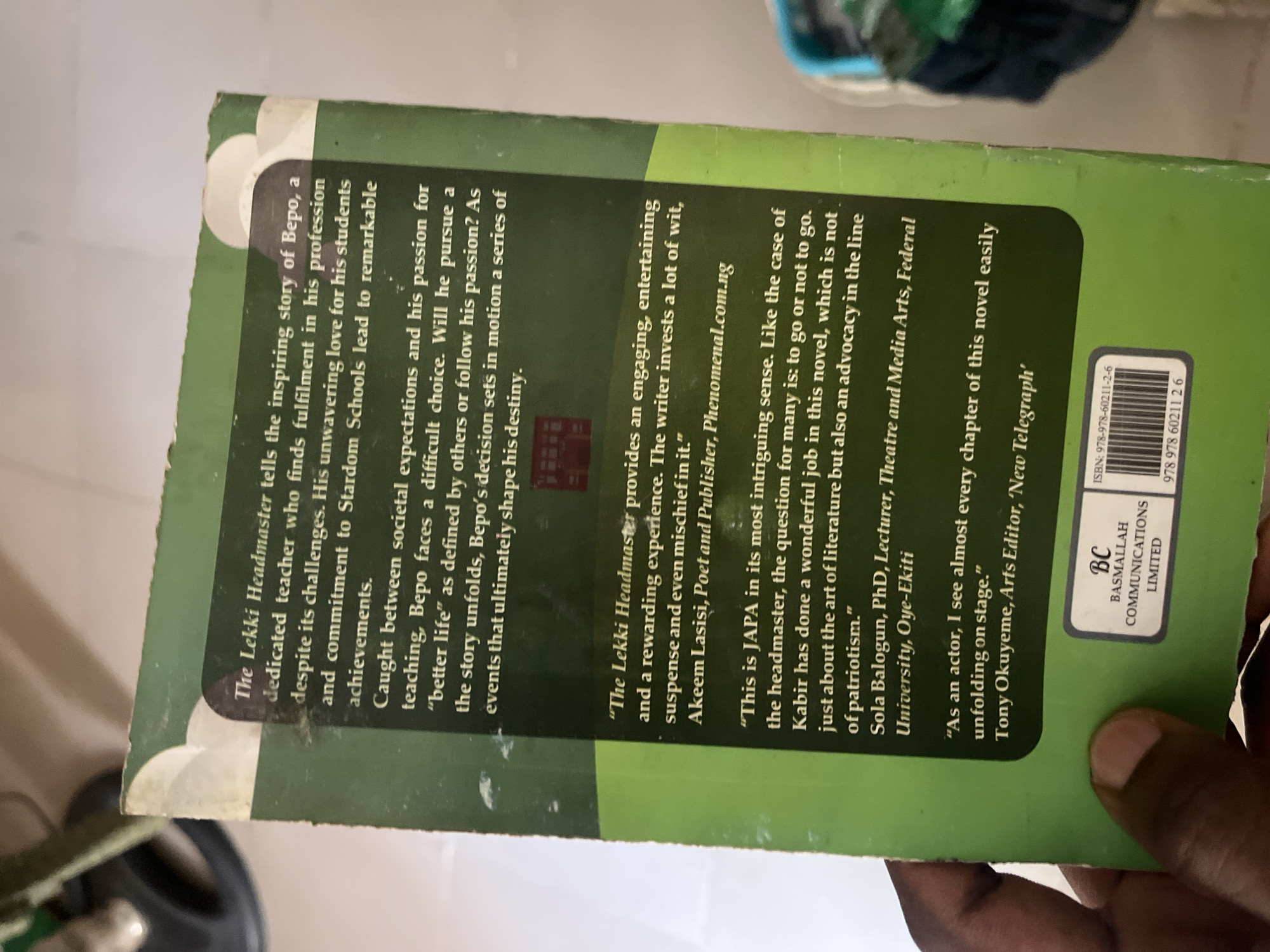 The image shows the back cover of a book titled "The Lekki Headmaster". The cover is predominantly green with white text and some graphic elements. The text provides a synopsis of the book, highlighting the story of Bepo, a dedicated teacher facing challenges and pursuing his passion. It also includes quotes from reviewers, identifying them as Akeem Lasisi, Poet and Publisher, Phenomenal.com.ng, and Sola Balogun, PhD, Lecturer, Theatre and Media Arts, Federal University, Oye-Ekiti. Towards the bottom, there is a logo for "BC BASMALLAH COMMUNICATIONS LIMITED" and an ISBN barcode with the number 978-978-60211-2-6. The book cover appears slightly worn, with minor scuff marks. The image is taken from a close-up perspective, with a person's hand visible in the bottom left corner, holding the book. The background is blurred and out of focus, suggesting an indoor setting. There is no visible indication of the city of Jalingo, Nigeria, in the image.