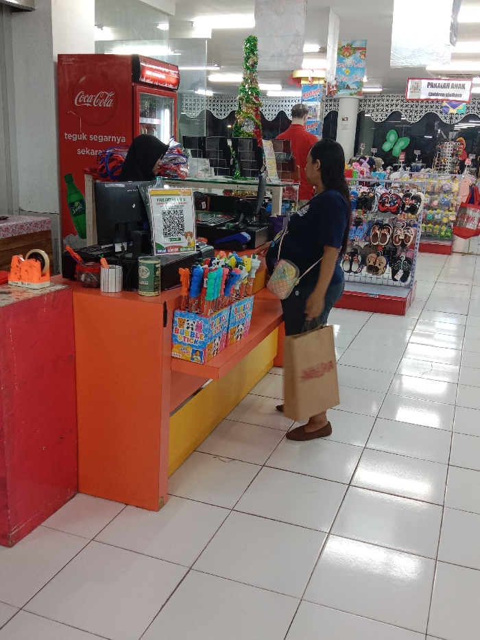 An interior shot depicts a retail store or supermarket checkout area in Langenharjo, Indonesia. A female cashier, wearing a black hijab, is partially visible behind an orange and yellow counter, attending to a transaction. A monitor and a QR code payment sign are present on the counter. In front of the counter, a female customer stands, holding a large brown paper bag. She is wearing a dark blue shirt, blue jeans, and brown flat shoes. To the left of the cashier, a red Coca-Cola branded refrigerator is visible. The background features various merchandise displays, including children's footwear and clothing, and holiday-themed decorations like a green tinsel-wrapped column and hanging signs. The flooring consists of white square tiles. A male figure in a red shirt is partially visible further in the background.