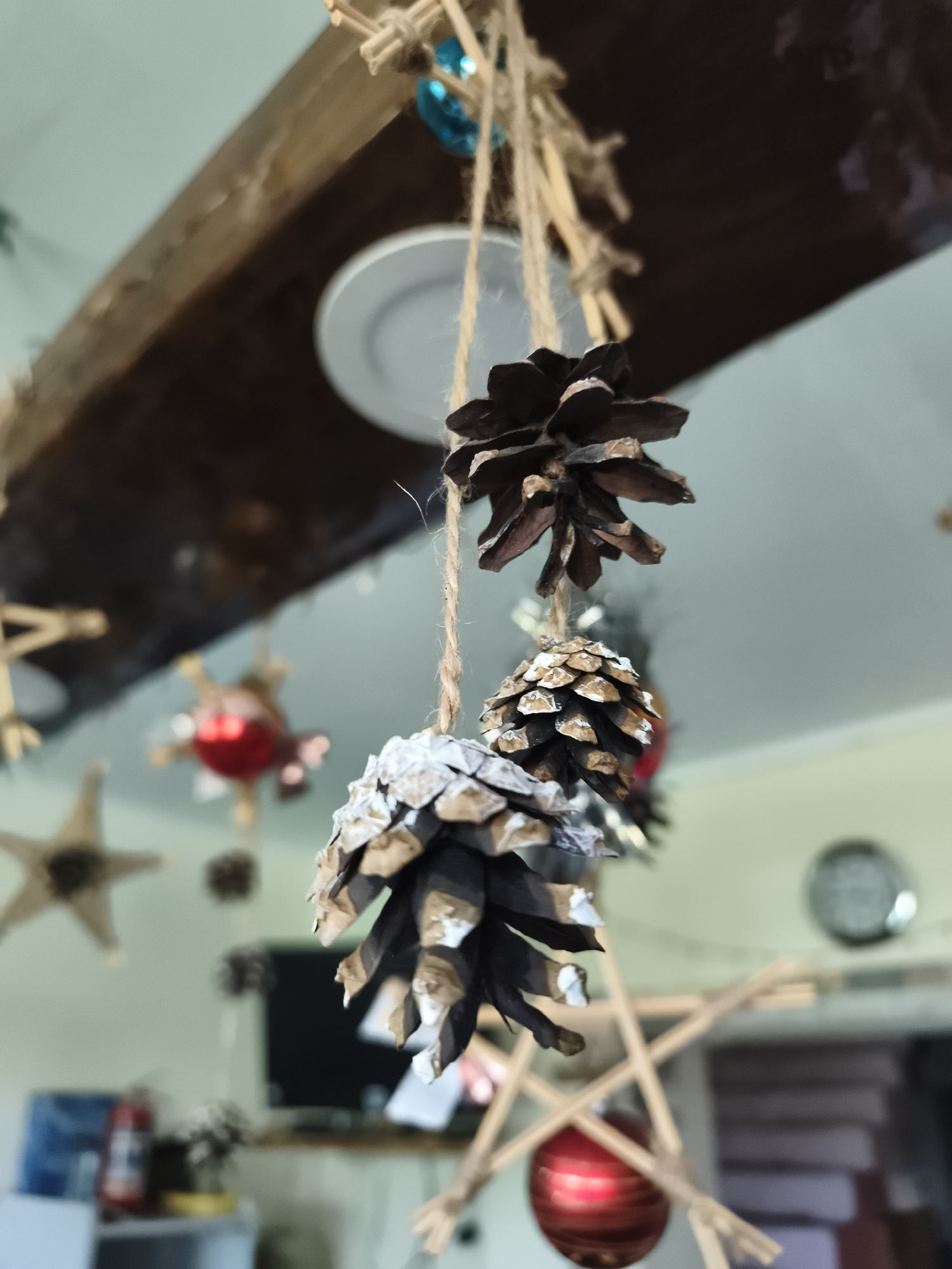 The image shows a close-up of pinecone decorations hanging from twine, suspended from a wooden beam. The pinecones are dark brown with some of their tips painted white, possibly to resemble snow. They are arranged in a vertical line, with the largest pinecone at the top and smaller ones below. In the background, out of focus, are various other decorations including a star shape made of twigs, a red spherical ornament, and what appears to be a clock on the wall. The environment suggests an indoor setting, likely a room with rustic or festive decor. There is no indication of the specific location within Гаранскі сельскі Савет, Belarus.