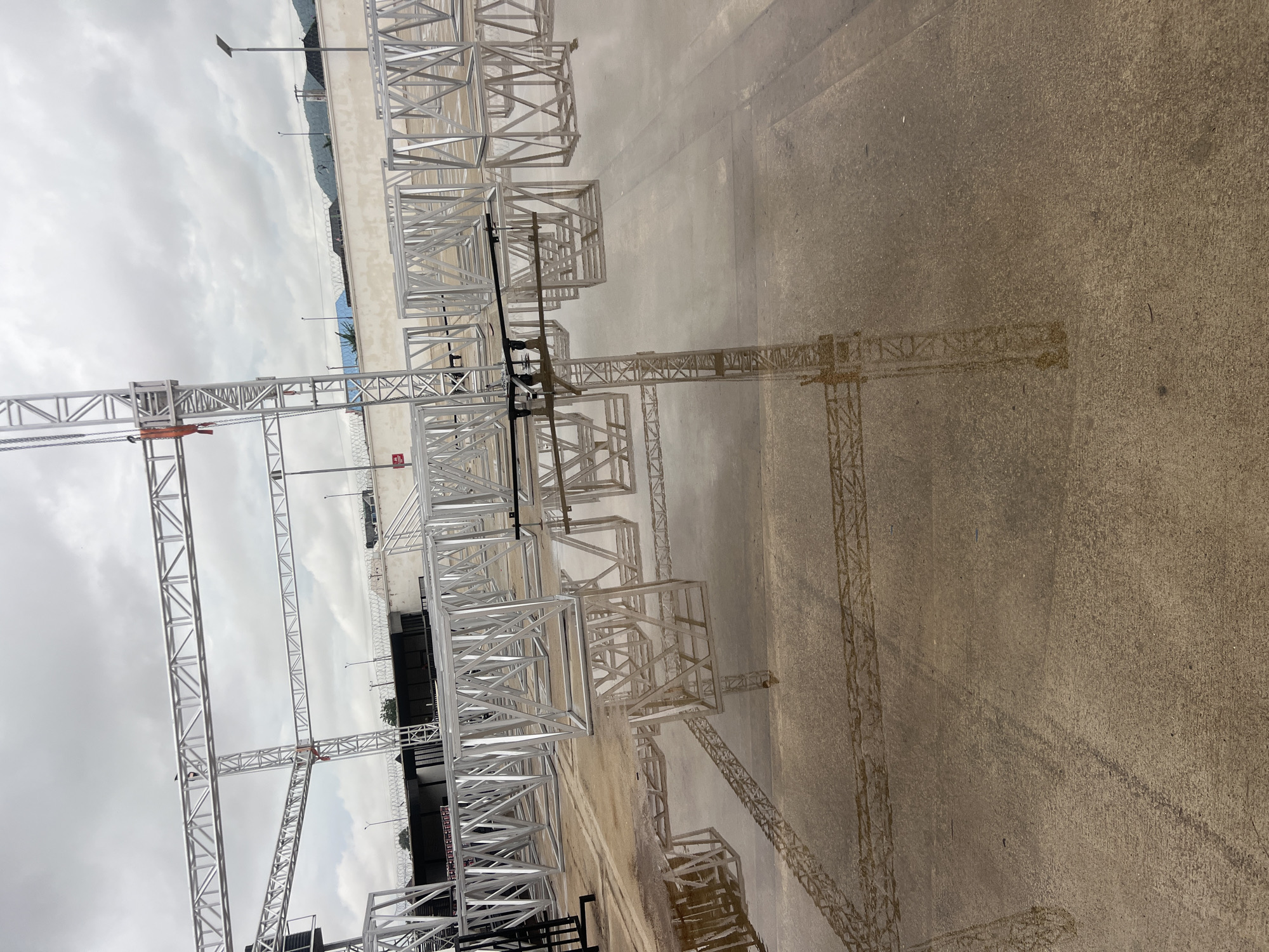 The image shows a collection of metal truss structures, some assembled into large frames and others stacked in modular units, on a wet concrete surface. The sky is overcast and the ground is covered in puddles reflecting the structures and the sky. In the background, a white wall with barbed wire is visible, behind which are some buildings and a blurred landscape of hills. The scene appears to be an outdoor staging or construction area, possibly for an event, in Howayi, Nigeria. There are no people visible in the image. The lighting suggests it is daytime.