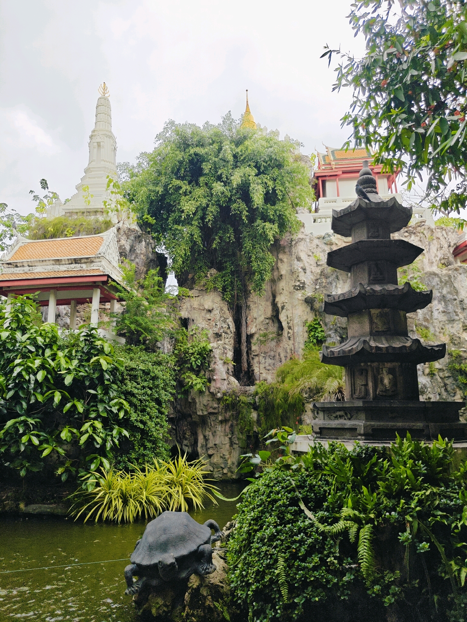 This image captures a serene scene at Wat Saket, also known as the Golden Mount, in Bangkok, Thailand. In the foreground, a dark stone sculpture of a turtle rests in a pond, surrounded by lush green foliage and some bright yellow plants. To the right, a multi-tiered dark stone pagoda stands tall, with a few birds perched on its top. Behind the pagoda and the pond, a rocky outcrop is visible, adorned with vegetation and a small temple-like structure with an orange tiled roof. Further in the background, a tall white stupa with a golden finial crowns the top of the rocky hill, and another building with a golden spire is also visible. The sky is overcast, contributing to the tranquil atmosphere of the scene.