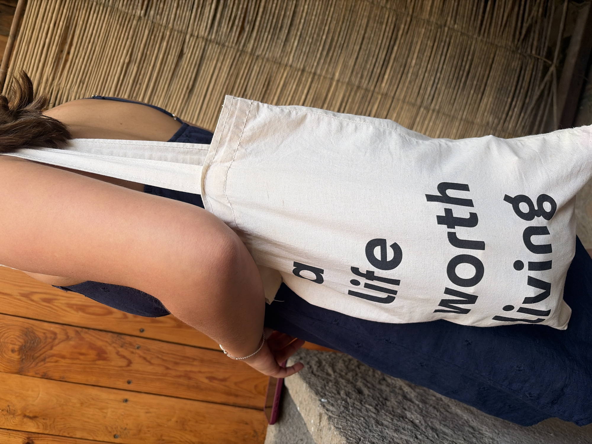 A person wearing a blue garment is seated on a wooden surface, next to a concrete step. They are holding a white tote bag with black text that reads "a life worth living." The background consists of a woven wall.