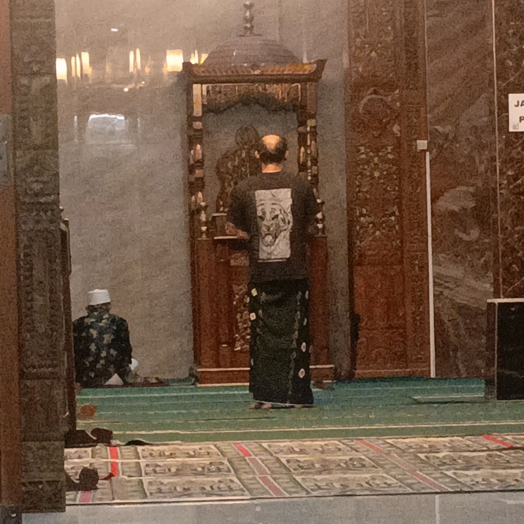The image depicts the interior of a mosque in Salatiga, Indonesia. Two individuals are present. In the foreground, a person is kneeling on a green carpet with white lines, facing away from the camera. This individual wears a white skullcap and a patterned garment. Further back, another person stands facing a decorative wooden structure, also with their back to the camera. This person is wearing a dark t-shirt with a graphic print and a patterned sarong. The mosque interior features ornate wooden carvings, lighting fixtures, and patterned prayer mats on the floor. The overall lighting suggests it might be evening or an interior with ambient lighting.