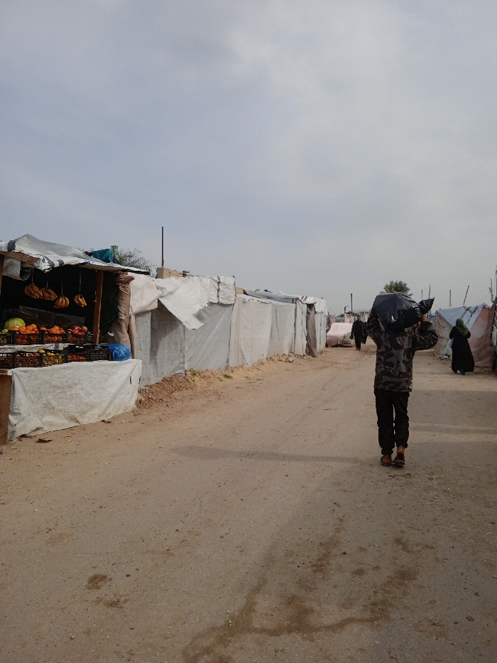 The image depicts an unpaved, dusty road within a makeshift settlement, identified as a location in Khan Yunis, Palestinian Territory, under an overcast sky. In the foreground, on the left, a rudimentary fruit stand is constructed from wooden poles and white tarps. It displays various fruits in black plastic crates, including oranges, apples, and bananas hanging from the canopy. A blue plastic bag rests on the stand. Extending beyond the stand along the left side of the road are numerous shelters, haphazardly constructed from white tarps and other materials, suggesting temporary or informal housing.
In the mid-ground, a person, facing away from the viewer, walks down the center-right of the road. This individual wears a camouflage-patterned long-sleeved top, dark trousers, and light-brown shoes, carrying a large, dark-colored plastic bag over their right shoulder and head. Further down the road, another person in dark clothing is visible walking in the same direction. On the far right, near the distant shelters, a third individual wearing a dark abaya and head covering also walks away. The road surface consists of compacted earth and sand with visible darker stains. The horizon reveals more such tarp-covered structures and a distant palm tree under a light grey, cloudy sky.
