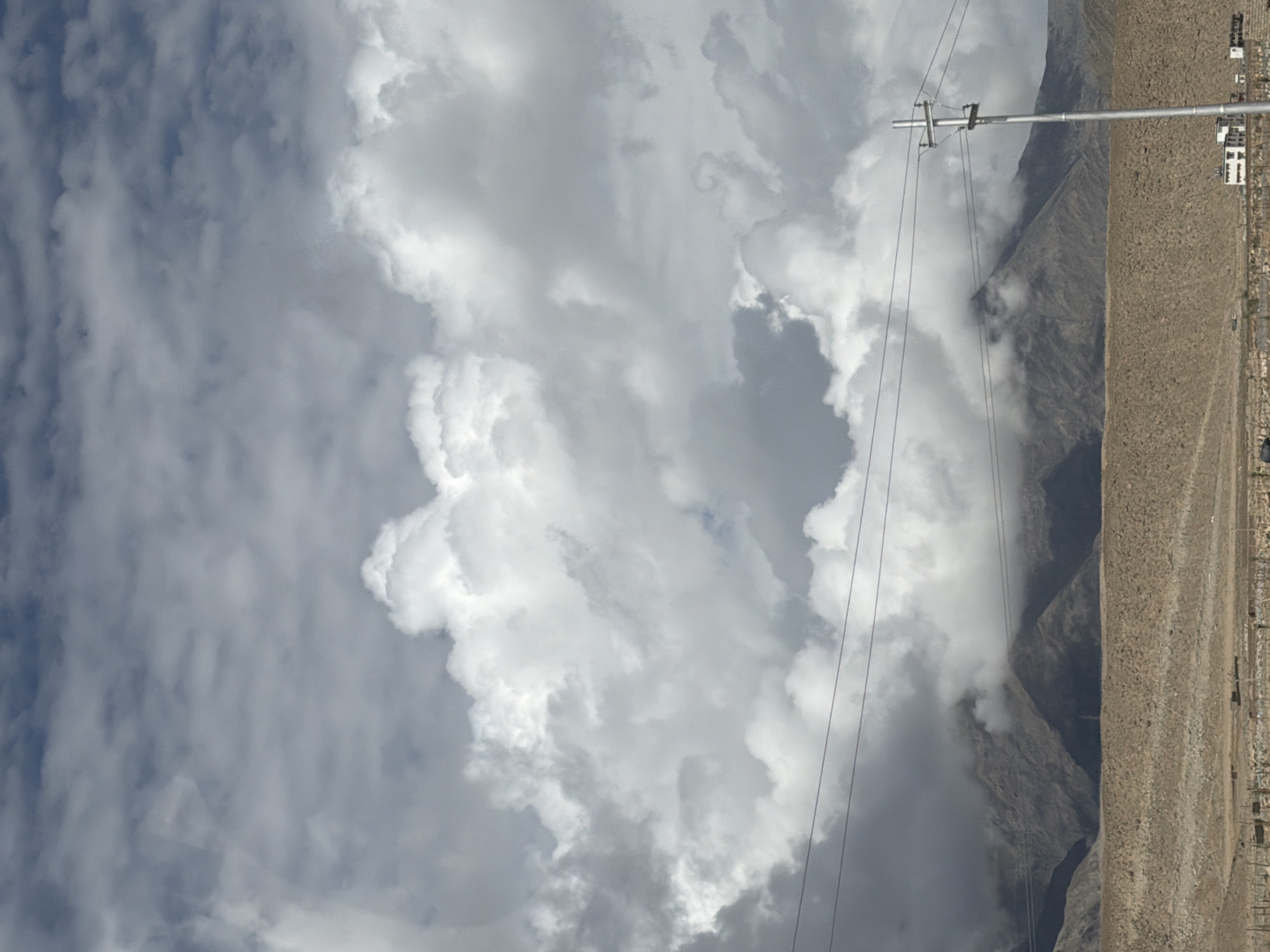 The image shows a wide shot of the mountainous terrain and sky near Chumkar, India. The sky is filled with dramatic, heavy clouds, with lighter, brighter clouds scattered among them. In the mid-ground, a mountain range is visible, partially obscured by clouds and mist. A dry, arid landscape stretches across the foreground, with sparse vegetation. A power line with poles cuts across the frame horizontally. To the right, a single, modern-looking building with white walls and dark windows is visible at the base of the mountains, suggesting a small settlement. There are no people or animals visible in the scene. The overall impression is one of a vast, rugged, and somewhat overcast natural environment.