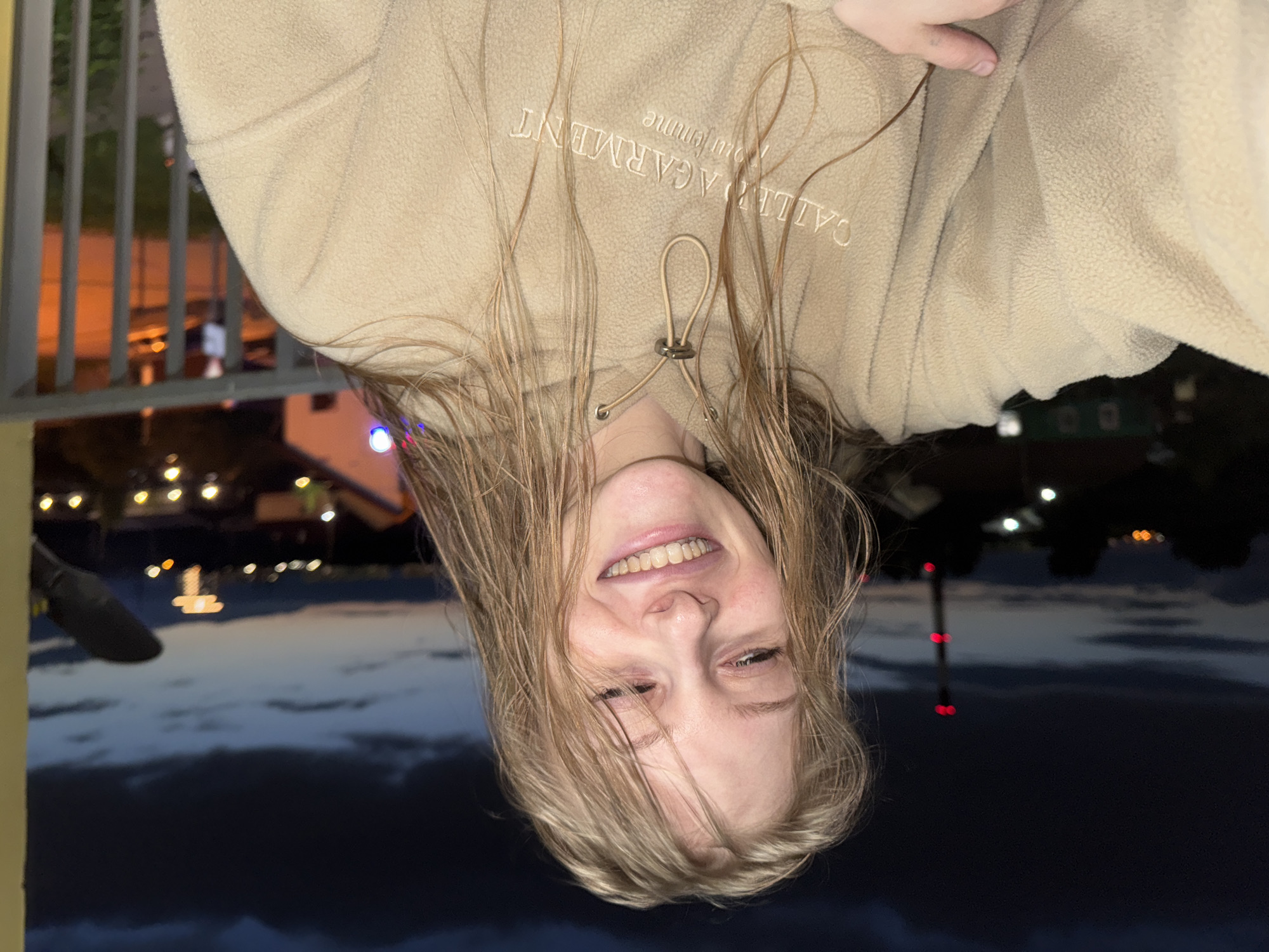 A young woman with blonde hair is pictured from the chest up, smiling and looking slightly down. She is wearing a tan hoodie with the embroidered text "CALLIOPE GARMENT" and "pour femme". Her hair is damp and falls around her face and shoulders. The background is a dark twilight sky with some clouds. In the distance, lights from buildings and a red-lit tower are visible, suggesting an urban or semi-urban environment. The woman is standing on a balcony or porch, indicated by a gray metal railing to her right. The overall scene suggests a casual, possibly outdoor moment at dusk. The location appears to be within or near Kondratovo, Russia, given the visible features of a populated area at night.