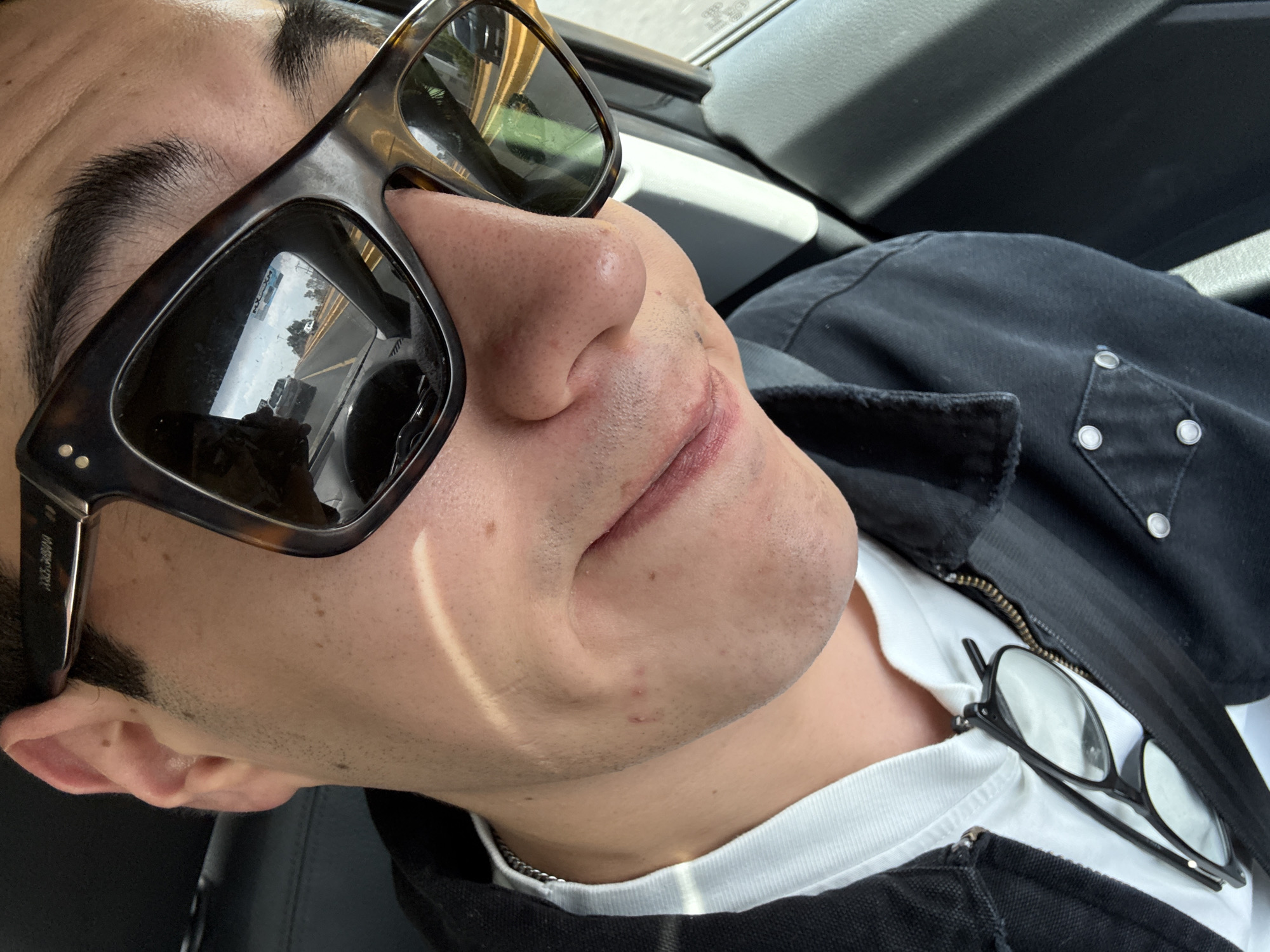 A close-up, canted shot depicts a person, presumably male, wearing tortoiseshell sunglasses, a white t-shirt, and a black jacket with snap closures on the pocket flaps. A second pair of eyeglasses rests on the t-shirt. The reflection in the sunglasses lenses shows an urban street scene, suggesting the subject is likely inside a vehicle. Based on the reflection within the sunglasses, the location is believed to be Puebla, Mexico. The person's expression is neutral, and the lighting is natural. The overall environment suggests being inside a car during daylight hours.