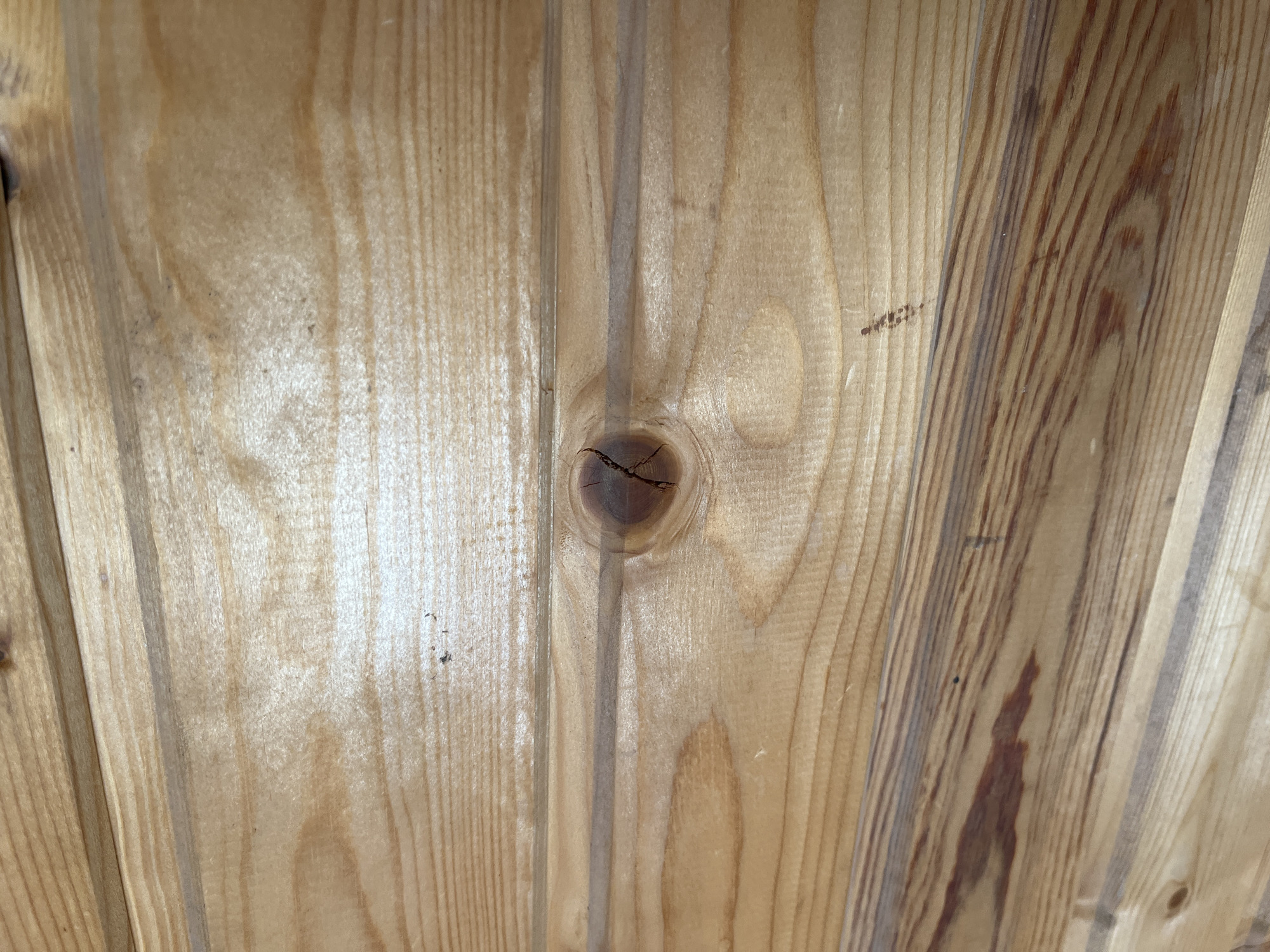 The image shows a close-up view of wooden paneling. The wood is light brown with visible grain patterns and knots. There is a prominent knot with a crack in the center of the image. The paneling appears to be arranged horizontally, with subtle indentations between the planks. The lighting suggests a well-lit indoor environment, possibly a room or a wall. There are no people or any other objects visible in the image besides the wood. The image does not provide any information about the location in Saratov, Russia.