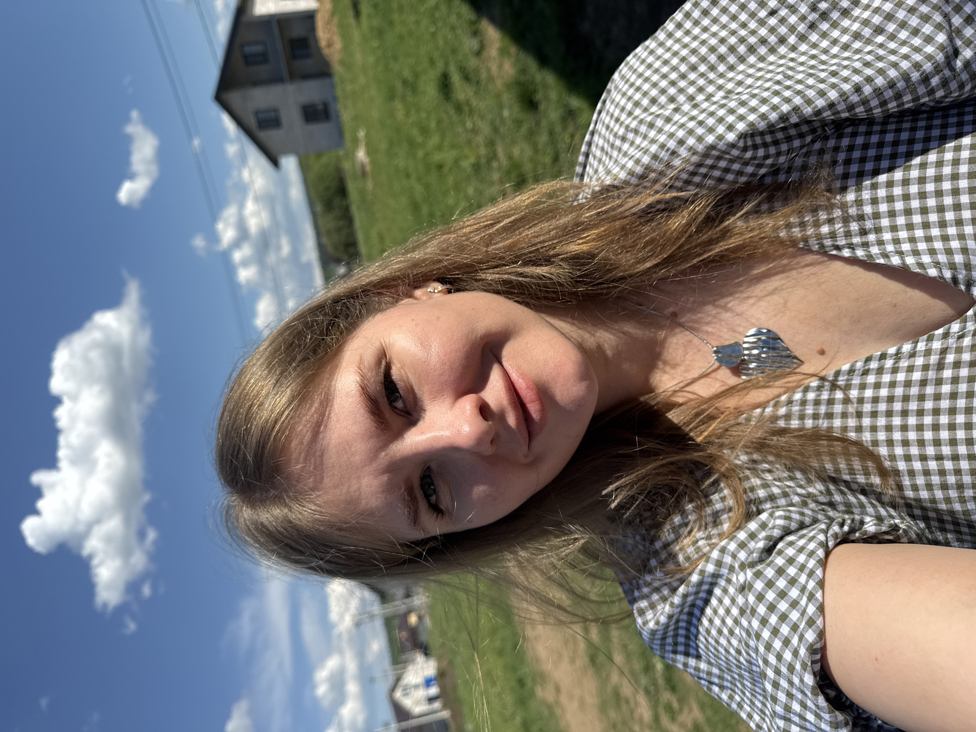 A woman is taking a selfie on a sunny day in Ферма, Russia. The woman is wearing a green and white checkered blouse and a silver heart necklace. The background includes green grass, a blue sky with clouds, and a building in the distance. Power lines are visible against the sky. Other buildings are also visible in the background.