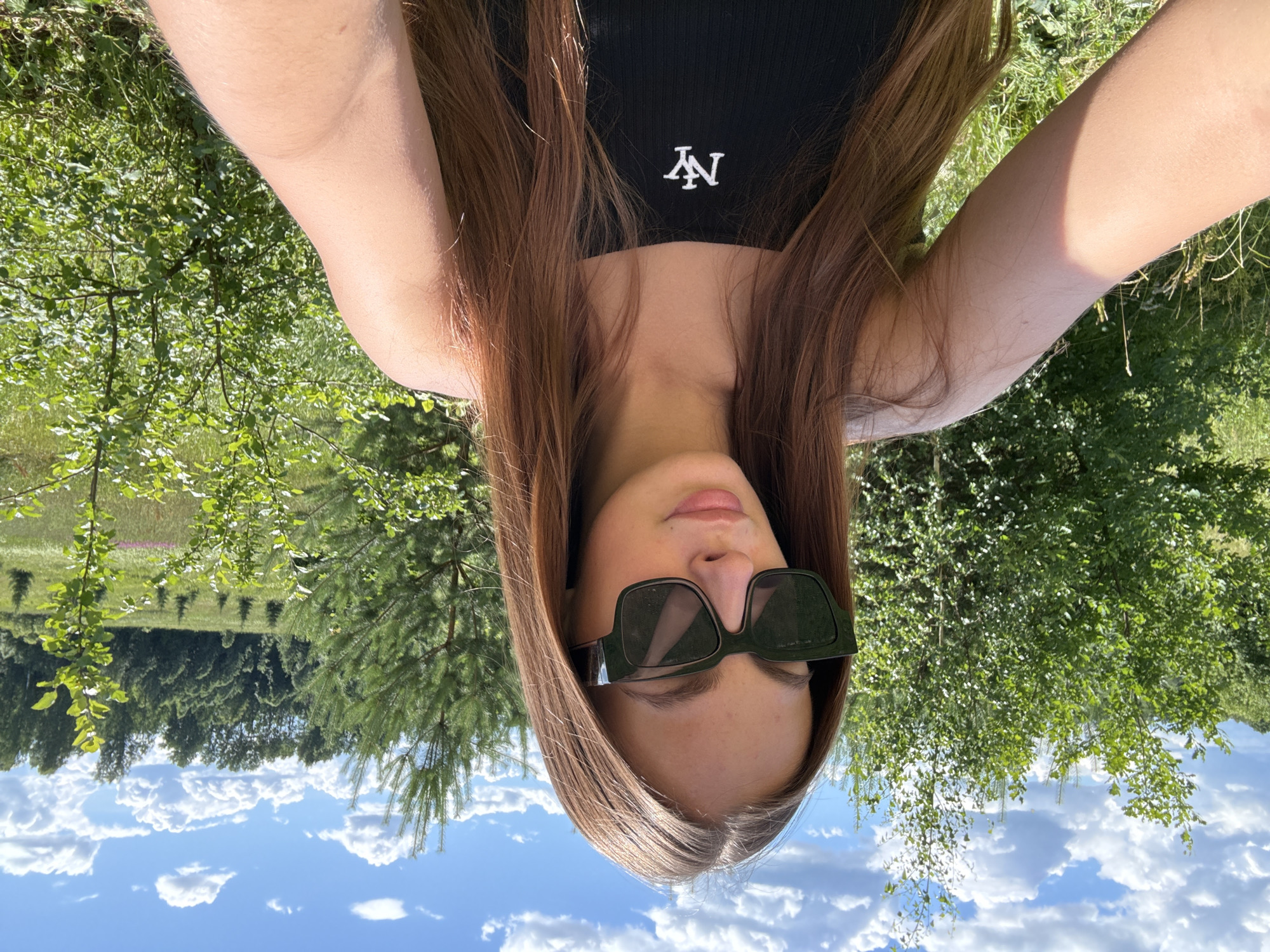 In Шолохово, Russia, a young woman with long, auburn hair, wearing dark sunglasses and a black tank top with a white "NY" logo, is taking a selfie outdoors. She is positioned in the foreground, with lush green foliage and trees forming the background. The sky is a bright blue with fluffy white clouds, suggesting a sunny day. The environment appears to be a natural, possibly rural or park-like setting, with various trees, bushes, and tall grass visible.