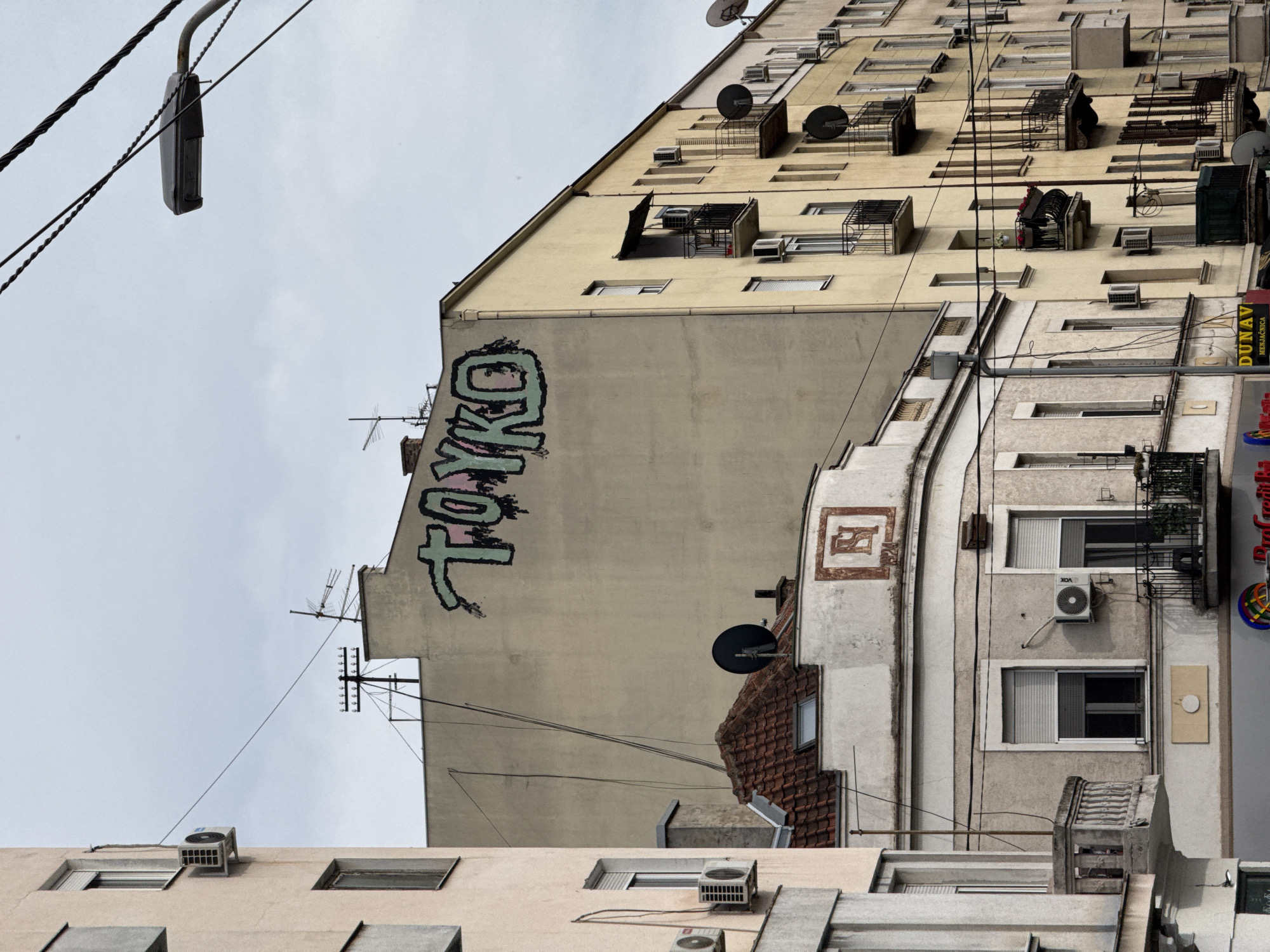 The image shows a street scene in Vracar Urban Municipality, Serbia. The view is angled upwards towards a cluster of buildings. One building has the word "TOKYO" in stylized graffiti on its facade. Another building has a repeating pattern of windows and air conditioning units. Satellite dishes are mounted on the roofs and sides of the buildings. Electrical wires crisscross the foreground of the sky.