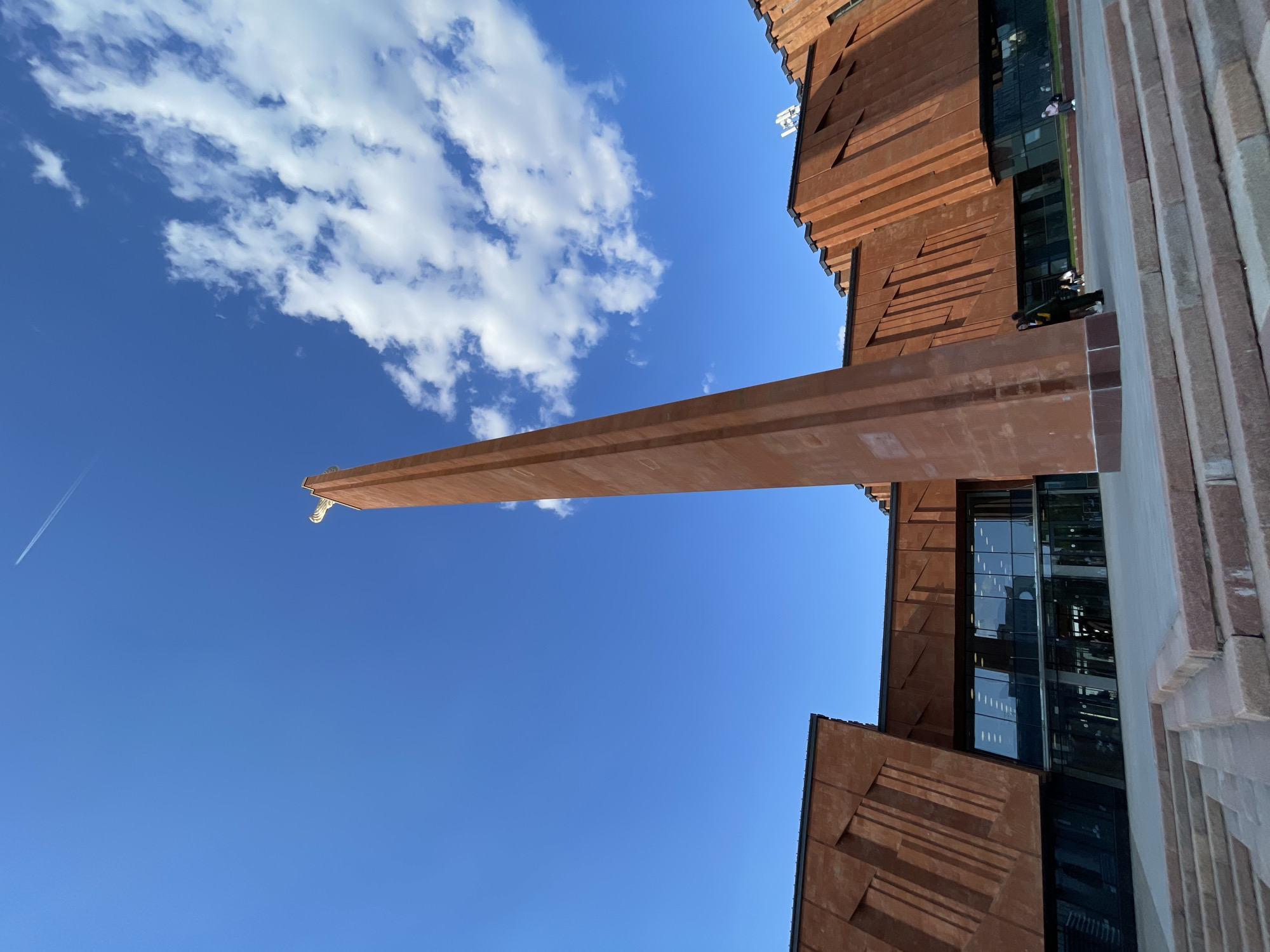 A low-angle, rotated image depicts the Kazan Family Center building in Kazan, Russia, against a bright blue sky with scattered white clouds. The building's exterior is clad in brown panels with a stepped design. A long, cantilevered extension juts out horizontally from the building. Glass windows reflect the sky. A few people are visible on the building's edge. A jet contrail cuts across the upper left sky.