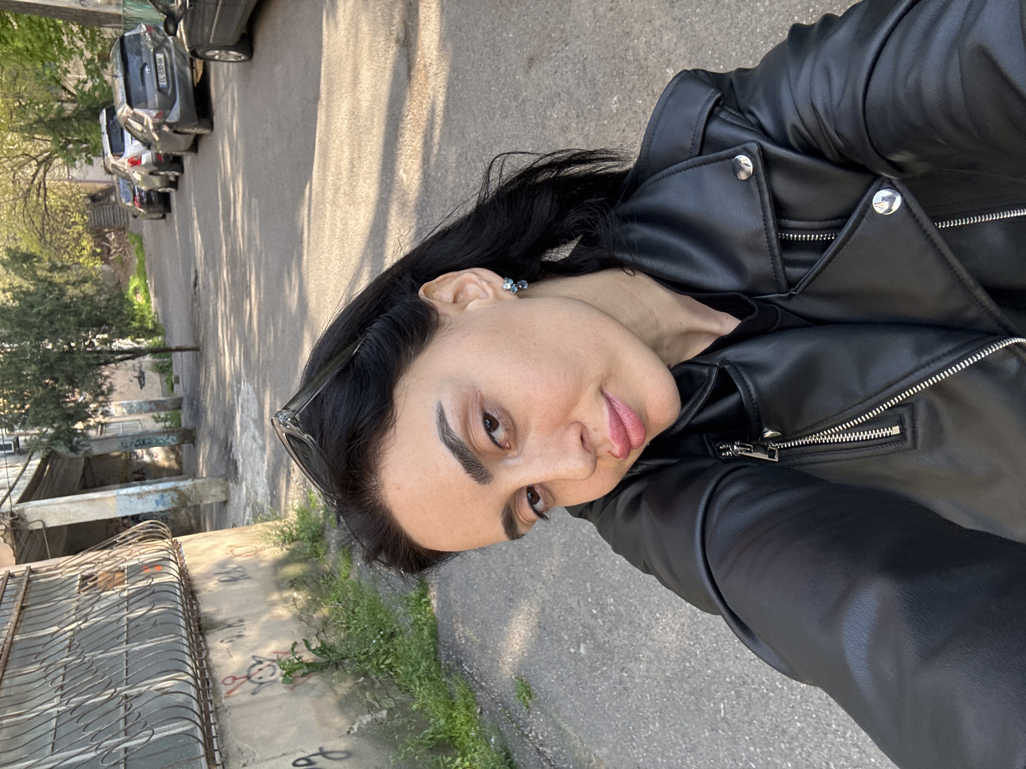 A woman in a black leather jacket is taking a selfie on a street in Tbilisi, Georgia. The street is lined with buildings, some of which are covered in graffiti. There are parked cars on the side of the road. The woman is wearing a silver earring.