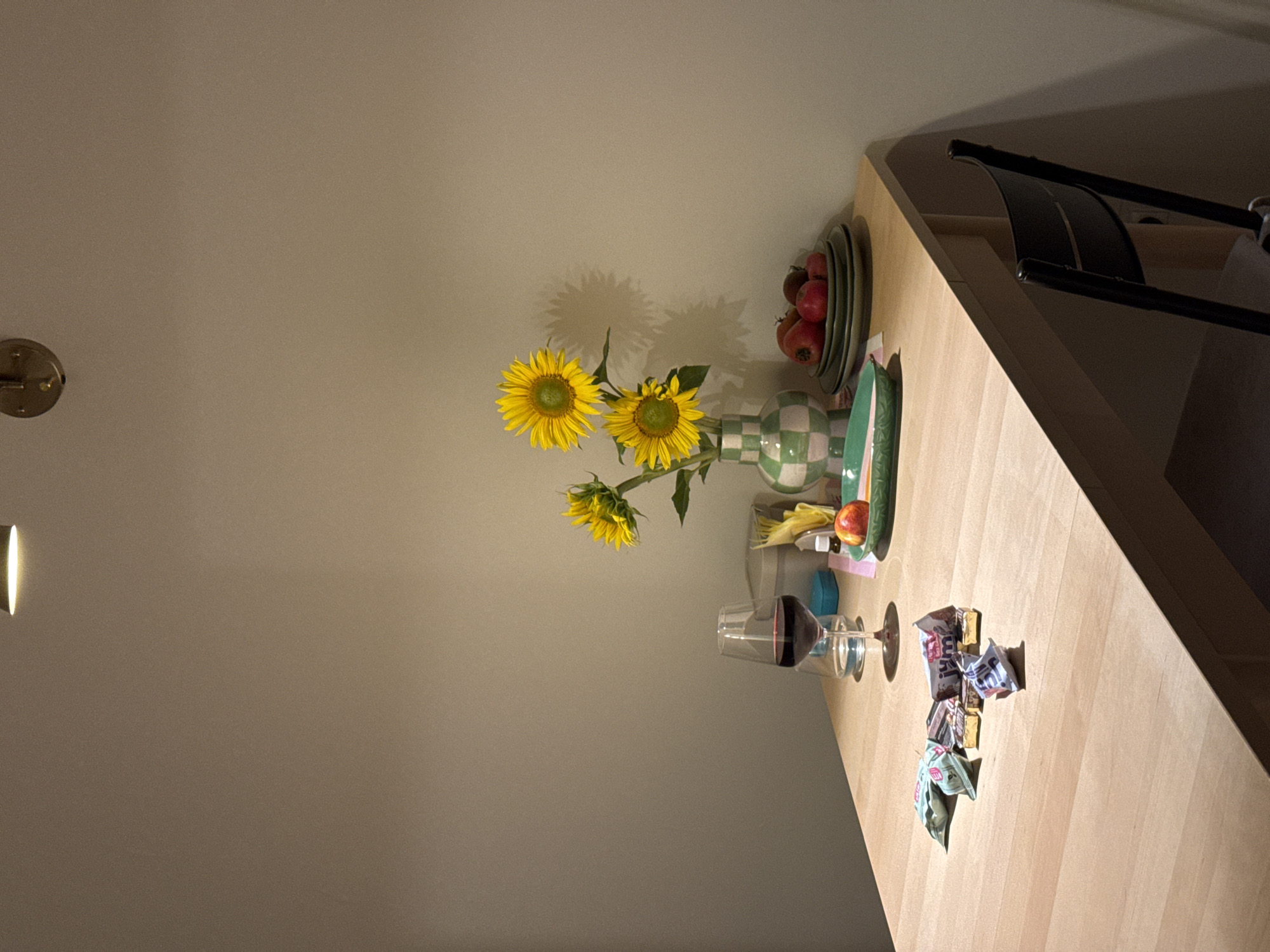 The image shows a wooden table set for a meal or snack. On the table, there is a glass of red wine, a small plate with an apple and some candies, a vase with sunflowers, and a stack of plates with tomatoes. A black chair is visible to the right of the table. The background is a plain wall. The lighting suggests an indoor setting, possibly a dining room or kitchen. The image does not provide any information that would allow for determining its location in Izhevsk, Russia.