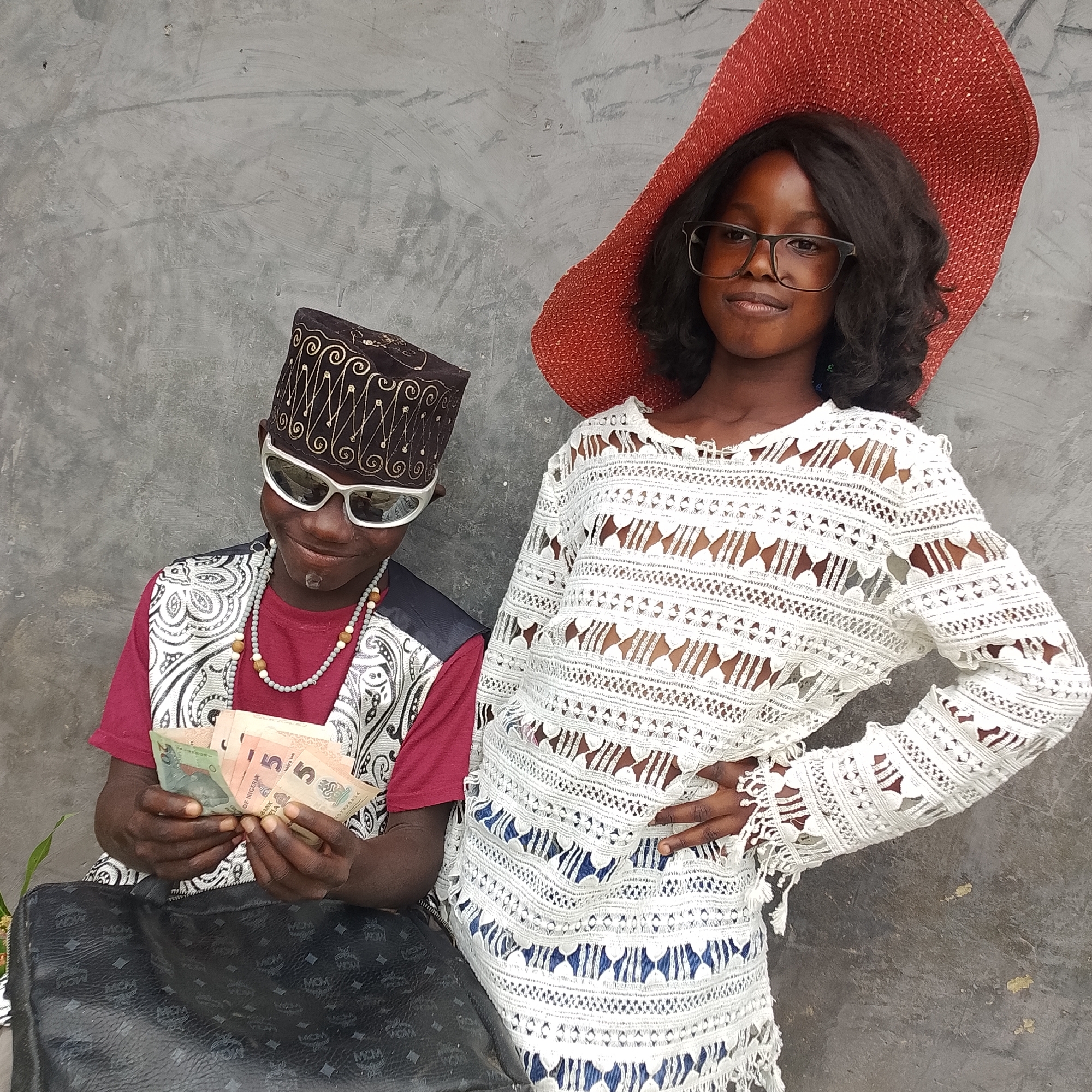 Two young people, a boy and a girl, pose against a rough, grey concrete wall. The boy, on the left, wears a traditional patterned cap, white sunglasses, a red t-shirt, and a vest with a black and white intricate pattern. He is holding several Nigerian Naira banknotes in his hands and appears to be counting them. A dark, patterned bag rests in front of him. The girl, on the right, wears a large, textured red sun hat and glasses. She is dressed in a white, lacy, long-sleeved top. Her left hand is on her hip. The scene appears to be outdoors. No specific location within Ado, Nigeria is discernible from the image.