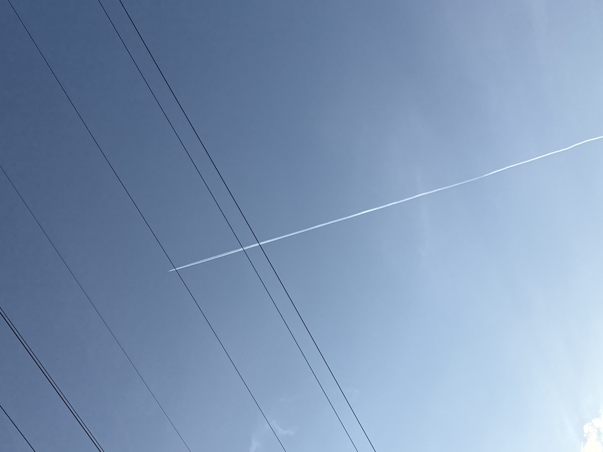 The image shows a clear blue sky with power lines running diagonally from the bottom left to the upper left. A contrail from an airplane cuts across the sky diagonally from the left to the upper right, appearing as a thin white line. The location is identified as Dolgoprudny, Russia. There are no people visible.