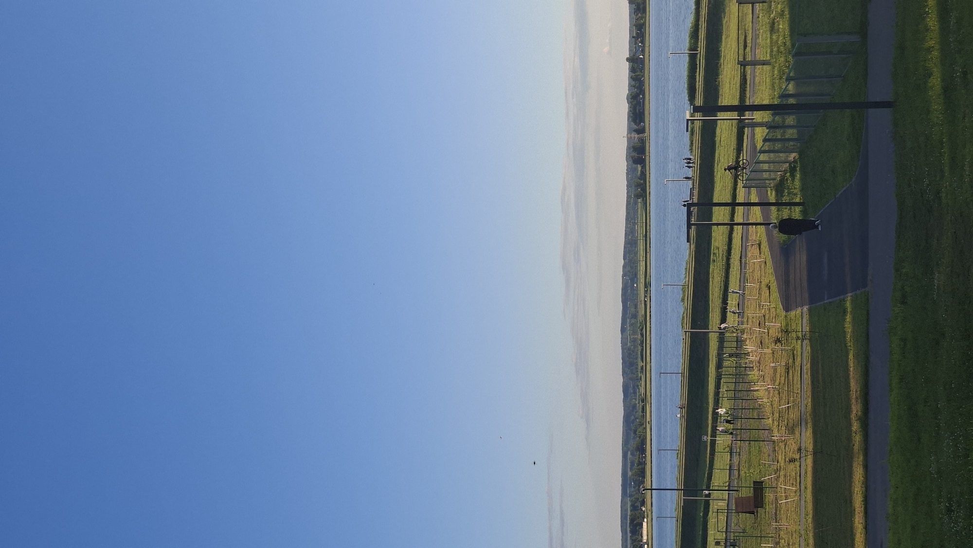 The image depicts a scenic view of the Zaslavsky Reservoir in Smylyavichy, Belarus. The reservoir dominates the mid-ground, reflecting the clear blue sky above. The shoreline is lined with a strip of green land, featuring a paved walkway curving through a park-like area. Several lampposts are positioned along the walkway and the perimeter of the park. In the foreground, a person walks along the paved path, and further back, other individuals can be seen strolling and one person is riding a bicycle. The distant background shows a low-lying, tree-covered landscape and scattered buildings, hinting at a town or village along the reservoir's edge. The overall atmosphere is peaceful and recreational.