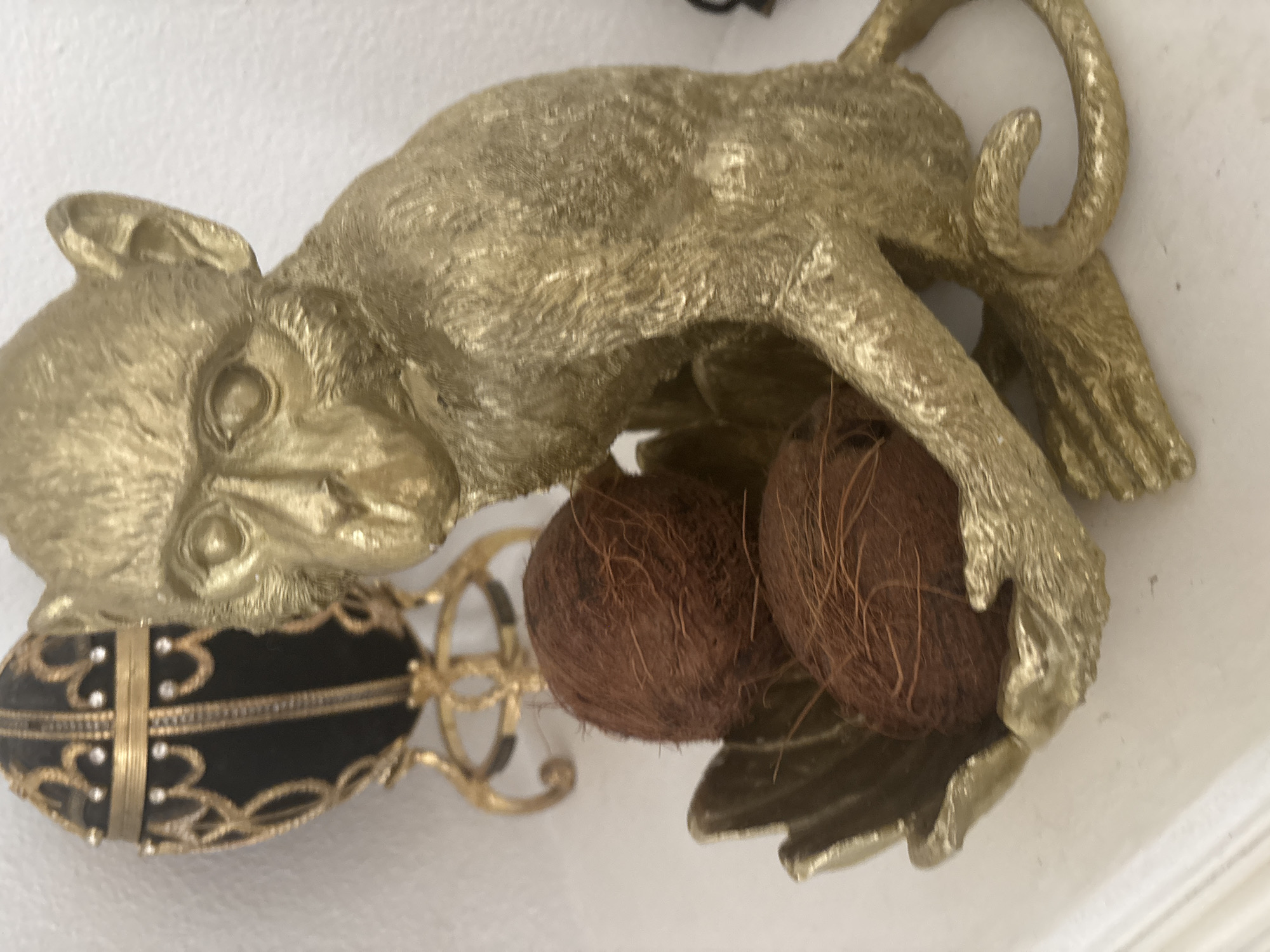 This is a close-up, eye-level, indoor shot of a decorative golden monkey figurine. The monkey is depicted in a crouching or lying-down position, with its body stretched out. It has a textured surface, suggesting fur, and is a bright, metallic gold color. The monkey's head is turned to its left, with its face clearly visible, featuring stylized eyes and ears.
The monkey is holding two coconuts in its arms or paws, which are nestled between its legs. The coconuts appear to be real, with their fibrous brown husks showing. To the left of the monkey, there is an ornate, oval-shaped object. This object is black with gold accents and appears to be decorated with small, clear stones, possibly diamonds or crystals. It is also embellished with intricate gold filigree and banding. The entire setup rests against a plain white background, likely a wall. The lighting appears to be soft and even, suggesting an indoor setting with artificial light. There is no visible text in the image.