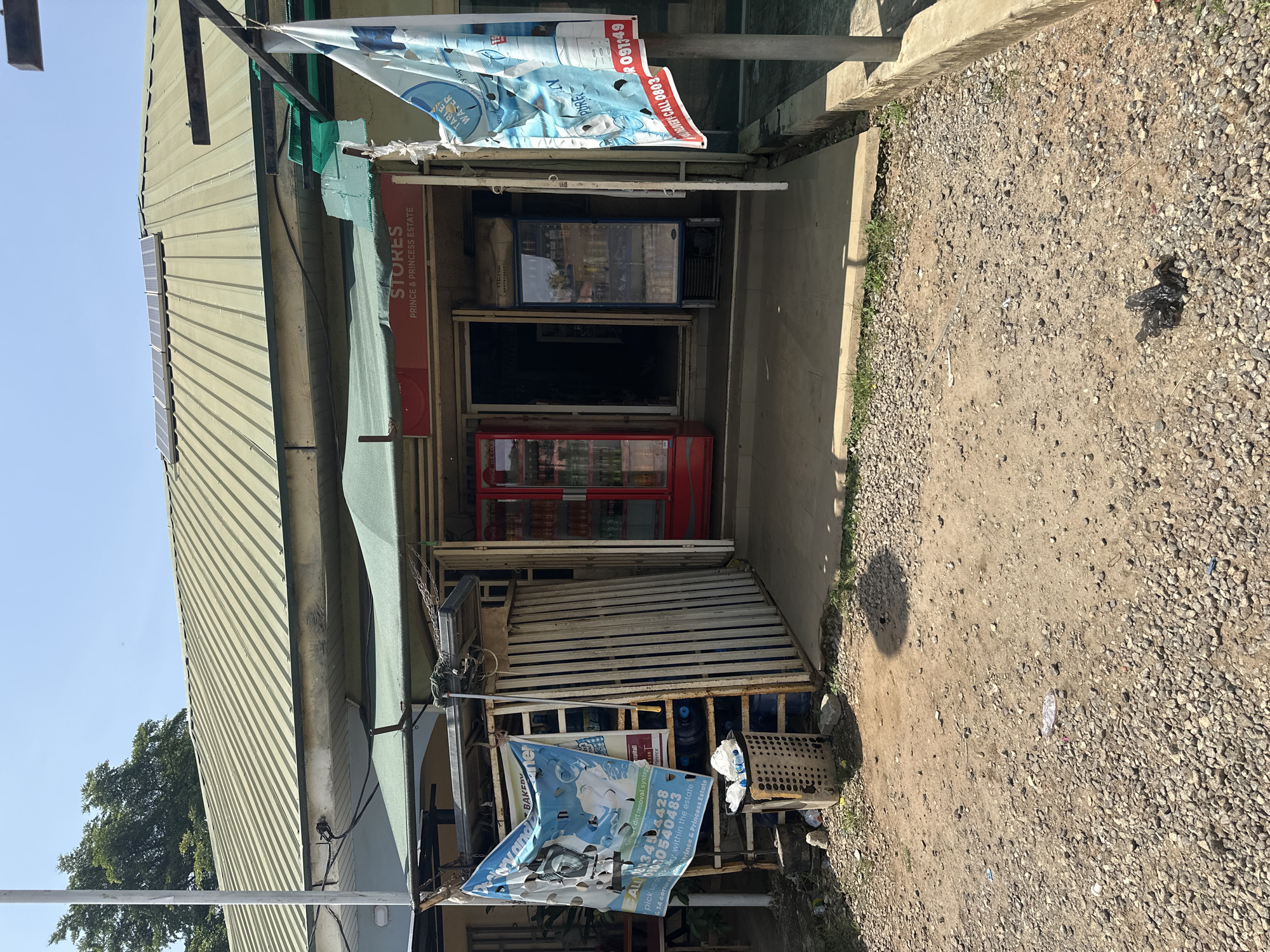 This is a photograph of a small convenience store in Abuja, Nigeria. The store appears to be open for business, with a red Coca-Cola refrigerator and a blue refrigerator visible inside. There are several advertisements displayed outside the store, including one for "PURE WATER" and another for "BAKERY." A sign above the entrance reads "STORES PRINCE & PRINCESS ESTATE." The ground in front of the store is covered in gravel, and there is a shadow cast on the ground, suggesting it is daytime. The building itself has a corrugated metal roof. There are no people clearly visible in the image, but the presence of a store suggests it is a place of commerce. The overall scene depicts a modest commercial establishment in a somewhat dusty environment.