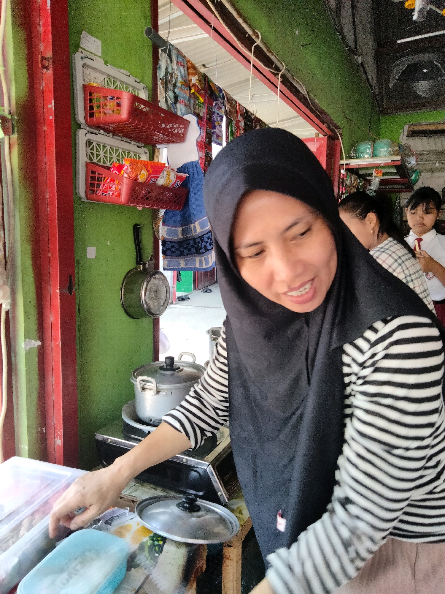 In Sidorejo, Indonesia, a woman wearing a black hijab and a black and white striped long-sleeved shirt is in the foreground, leaning over a table. She appears to be interacting with food items in clear plastic containers. Behind her, a second woman and a young girl in a school uniform are visible. The environment suggests a small shop or food stall with shelves displaying packaged snacks, cooking pots, and a hanging pan. The walls are painted green, and red trim is visible on a doorway.