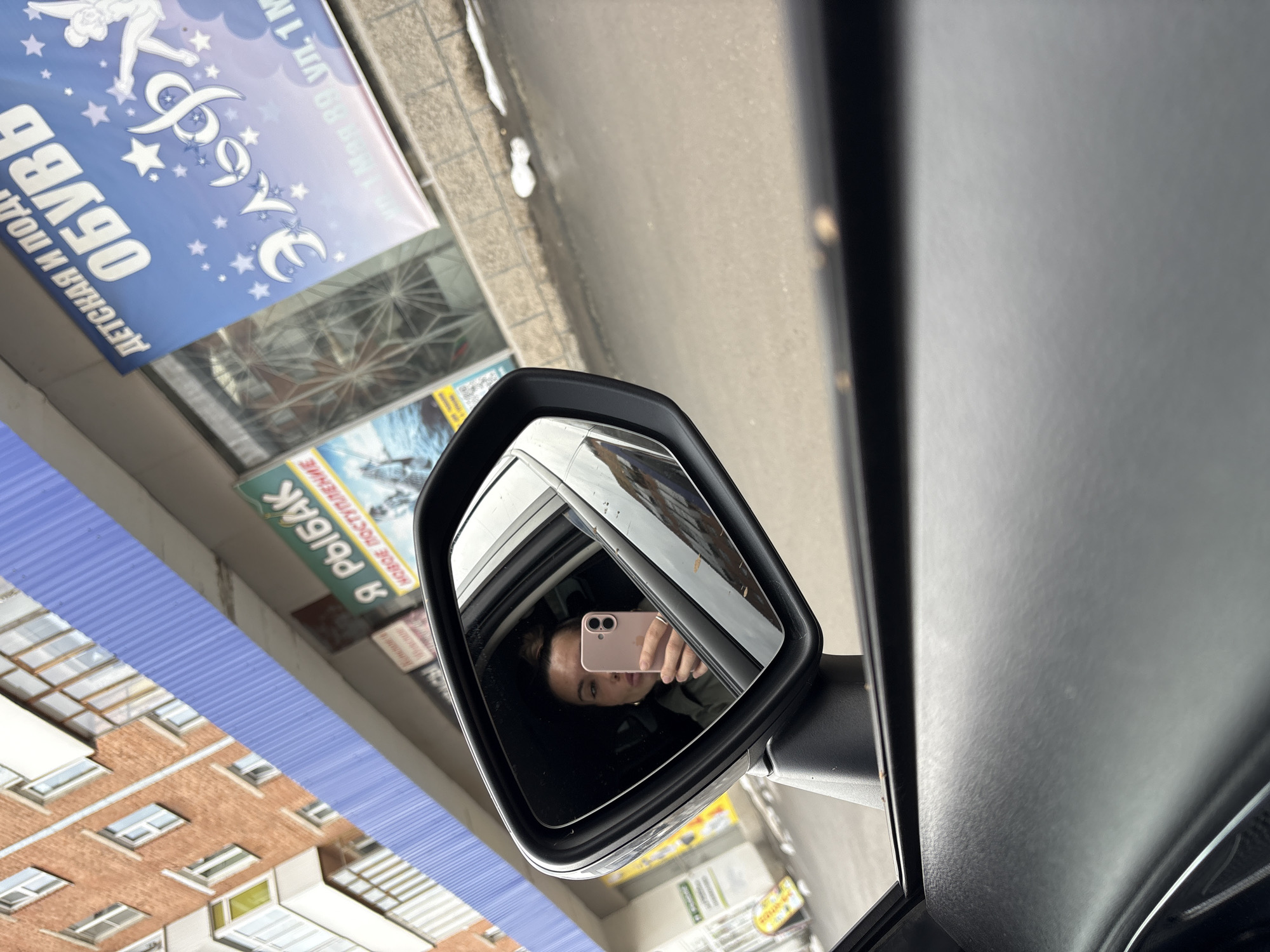 The image shows a side mirror reflection from inside a car. The reflection contains a woman taking a selfie with a pink phone. The background, as seen in the reflection, includes buildings and advertisements. The advertisements are written in Russian and include the words "ОБУВЬ" (shoes) and "Я РЫБАК" (I am a fisherman). One advertisement is on Ул. 1 Мая 89, Votkinsk, Russia. The time of day seems to be daytime due to the lighting conditions.