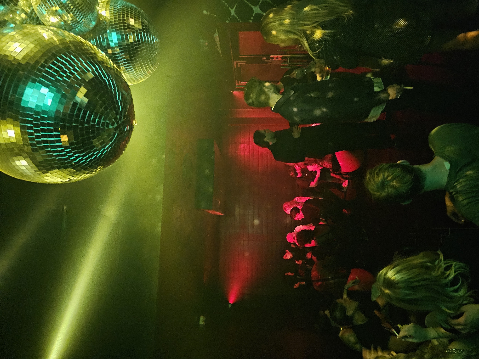 The image depicts a crowded nightclub scene, likely in Paris, France, given the style and ambiance. In the foreground, several disco balls reflect light, casting green and yellow hues across the room. A beam of light cuts through the smoky atmosphere. In the background, numerous people are gathered, illuminated by red and green stage lights. Some individuals are dressed in formal attire, including suits, suggesting a special event or upscale club. The crowd appears to be socializing and enjoying the music. The overall environment is dark, energetic, and celebratory.