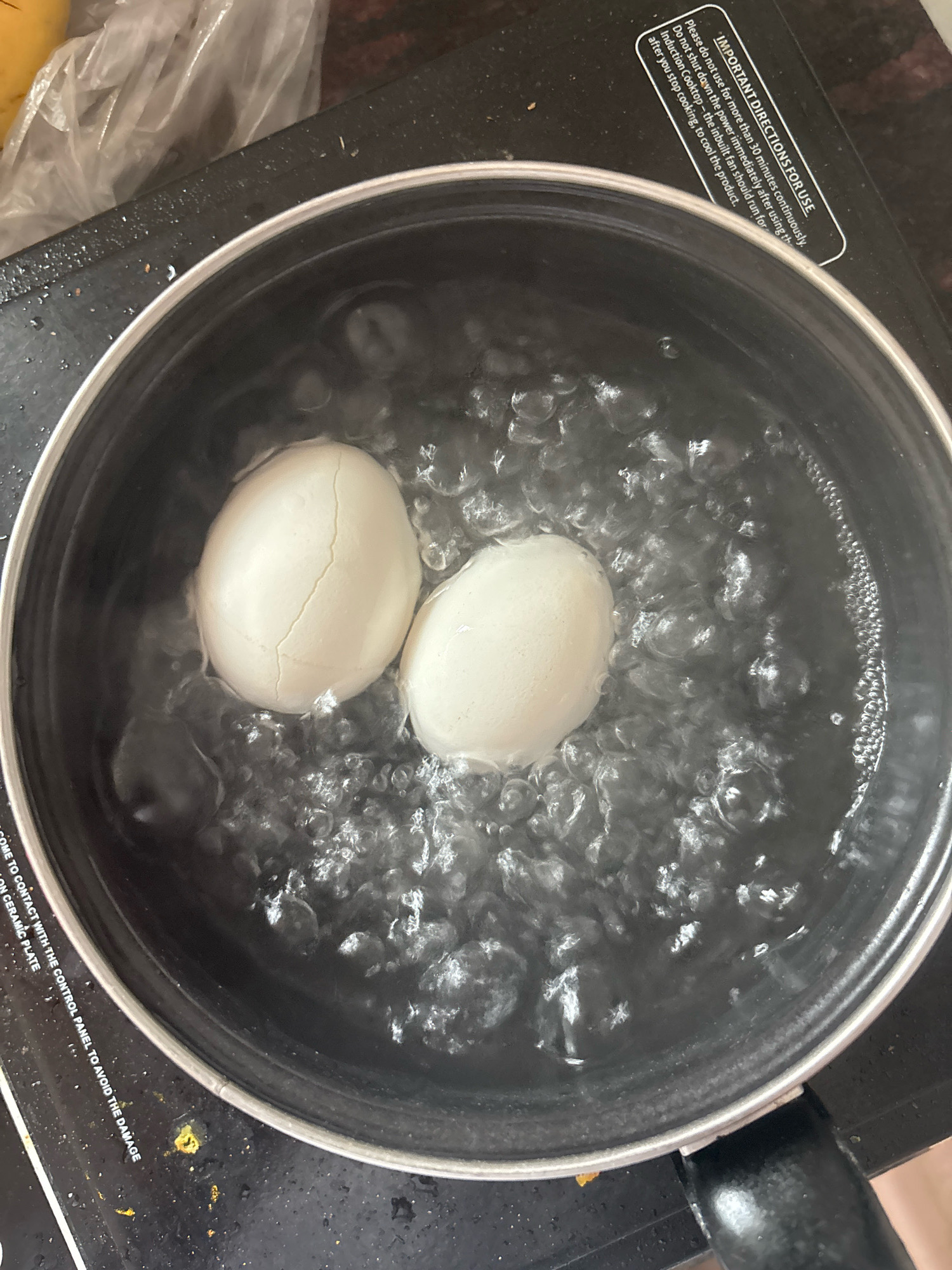 Two white eggs, one with a cracked shell, are submerged in boiling water in a stainless steel pot. The pot is placed on a black induction cooktop. Bubbles rise vigorously from the water around the eggs. The cooktop has white warning labels with black text. The environment appears to be a kitchen. There is no indication of the city of Bhatta, India, in the image.