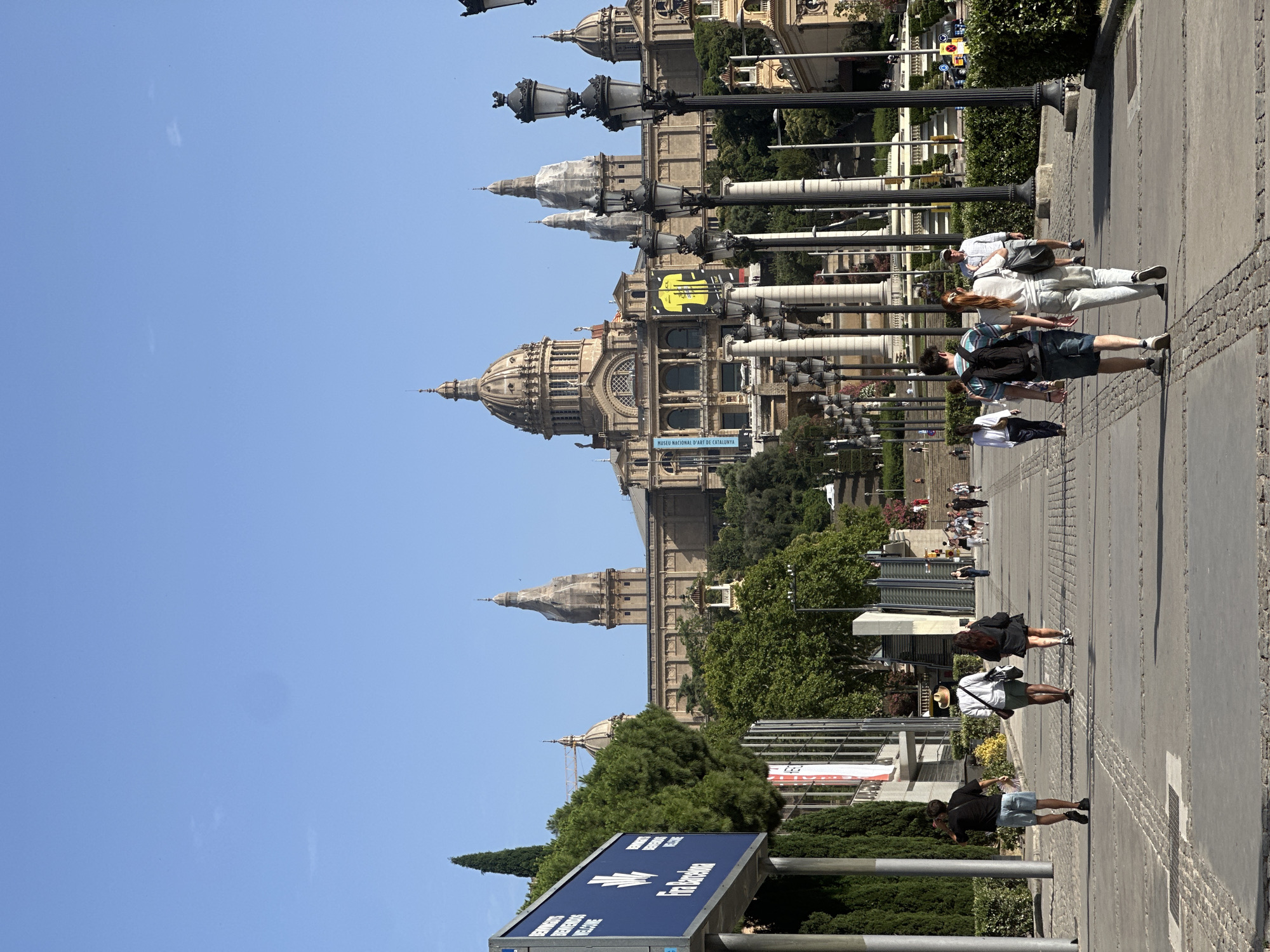 The image captures a daytime view of a wide, paved walkway leading towards the Palau Nacional, a grand building located on Montjuïc hill in Barcelona, Spain. The Palau Nacional features multiple domes and towers, with a prominent yellow banner displaying a football crest visible on its facade. A tall lamppost with multiple fixtures stands to the right of the walkway, framing the scene. Several individuals are walking along the path, some in small groups and others individually. They are dressed in casual summer attire. To the left of the walkway, a large blue sign with white lettering, possibly indicating an event or venue, is partially visible. Lush green trees and bushes line the sides of the walkway and surround the Palau Nacional, suggesting a park-like setting. The sky is clear and blue, indicating fair weather.